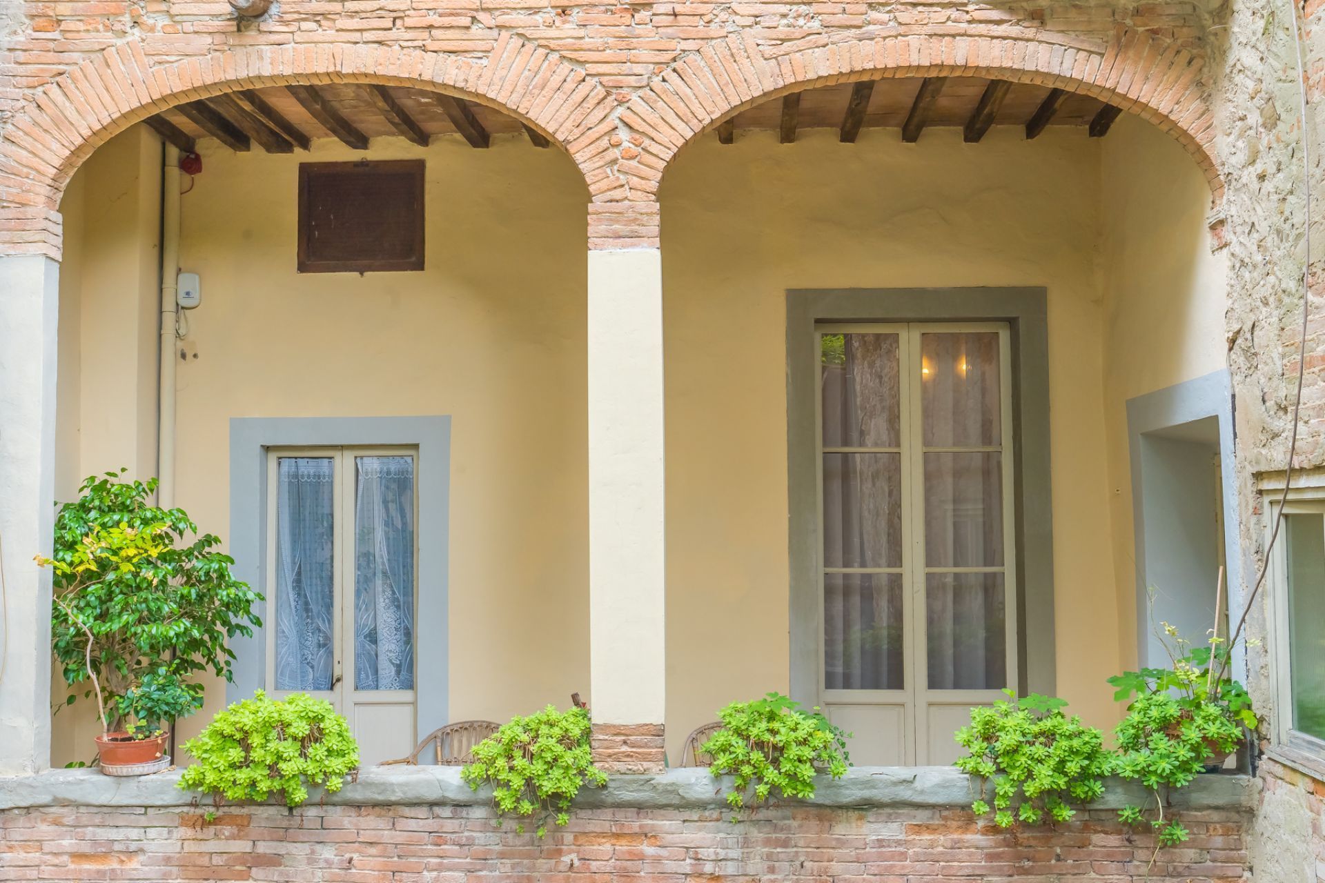 Balcone interno con aperture ad arco, pareti gialle, finiture in mattoni e arbusti in vaso lungo la ringhiera.