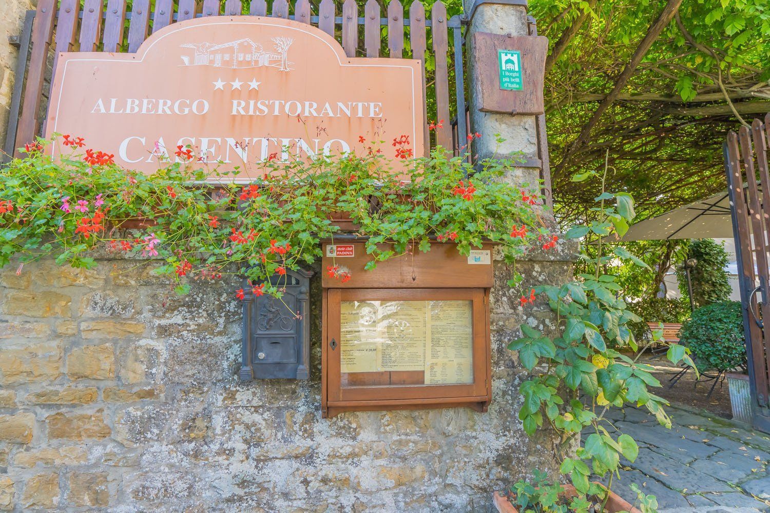 Ingresso rustico di un ristorante con insegna rosa, muro in pietra e fiori intorno a una vetrina in legno per il menù.
