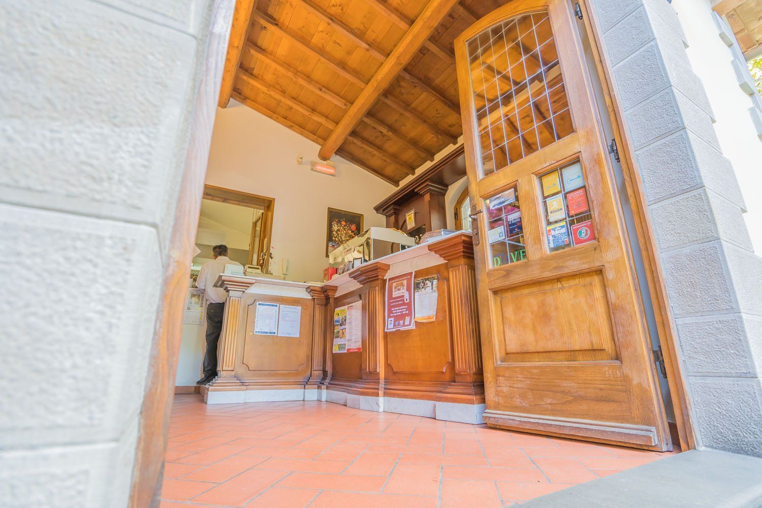 Bancone reception in legno all'interno di un ingresso piastrellato con porta aperta e soffitto alto con travi a vista.