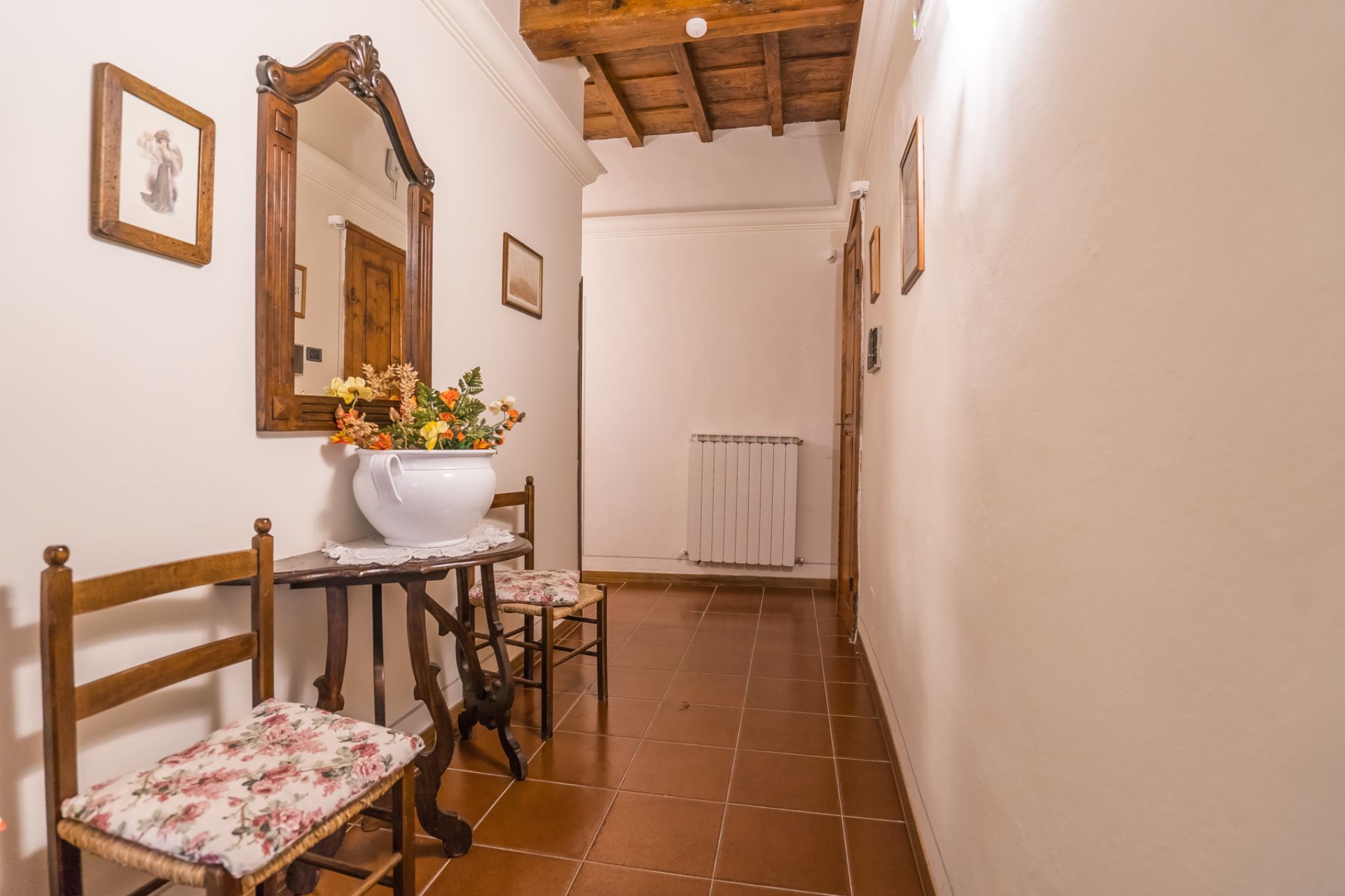 Stretto corridoio con pavimento in cotto, sedie in legno, specchio e un vaso di fiori.