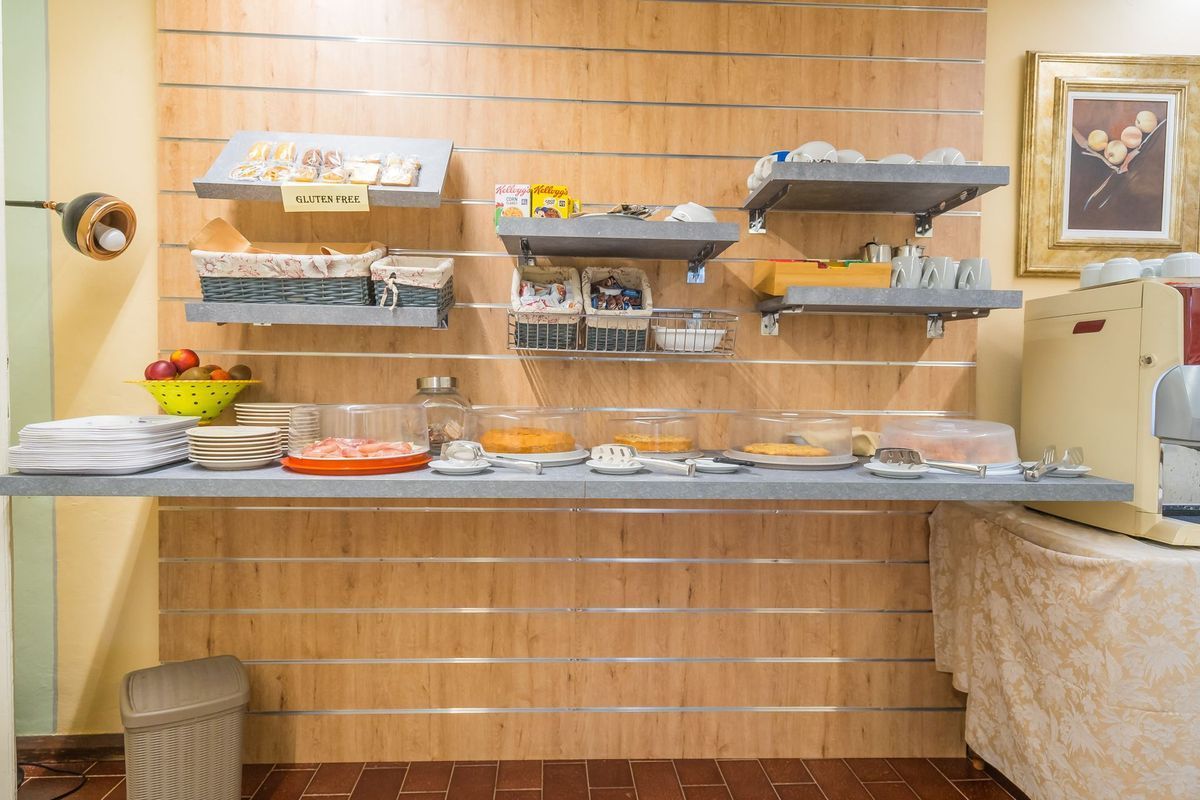 Colazione a buffet self-service con vassoi di cibo e piatti su un bancone nella zona pranzo.