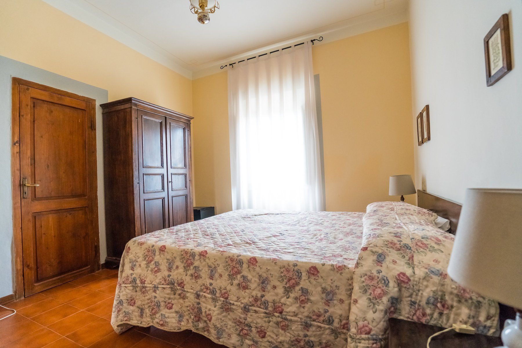Camera da letto soleggiata con copriletto floreale, armadio in legno e porta chiusa su pareti gialle