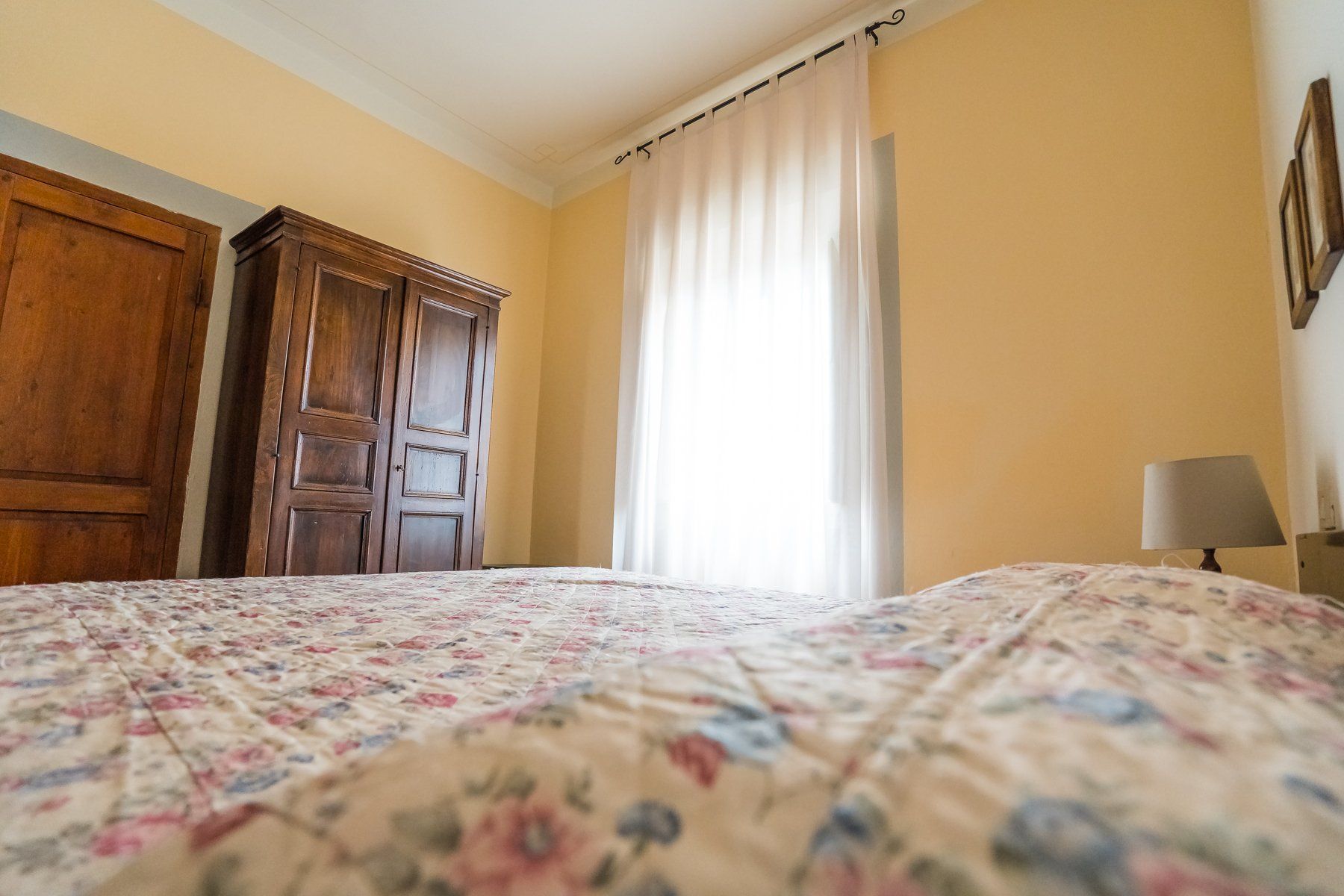 Camera da letto luminosa con copriletto floreale, armadio in legno e un'alta finestra con tende trasparenti.