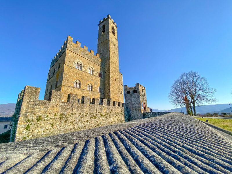 Castello di pietra con un'alta torre su un sentiero di ciottoli in pendenza sotto un cielo azzurro e limpido.