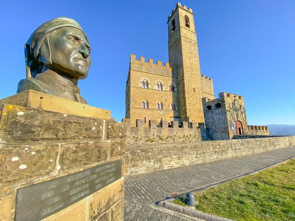 Busto di bronzo accanto a una torre di pietra su una collina soleggiata, con cielo azzurro e primo piano erboso.