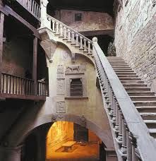 Scalinata storica in pietra con ringhiere decorate e arco illuminato da una luce calda in un cortile interno in penombra.