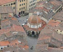 Veduta aerea di una piazza storica con una torre dell'orologio a cupola circondata da tetti di tegole rosse.