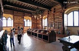 Persone che camminano in una sala museale rustica con pareti rivestite in legno, finestre ad arco e vetrine.