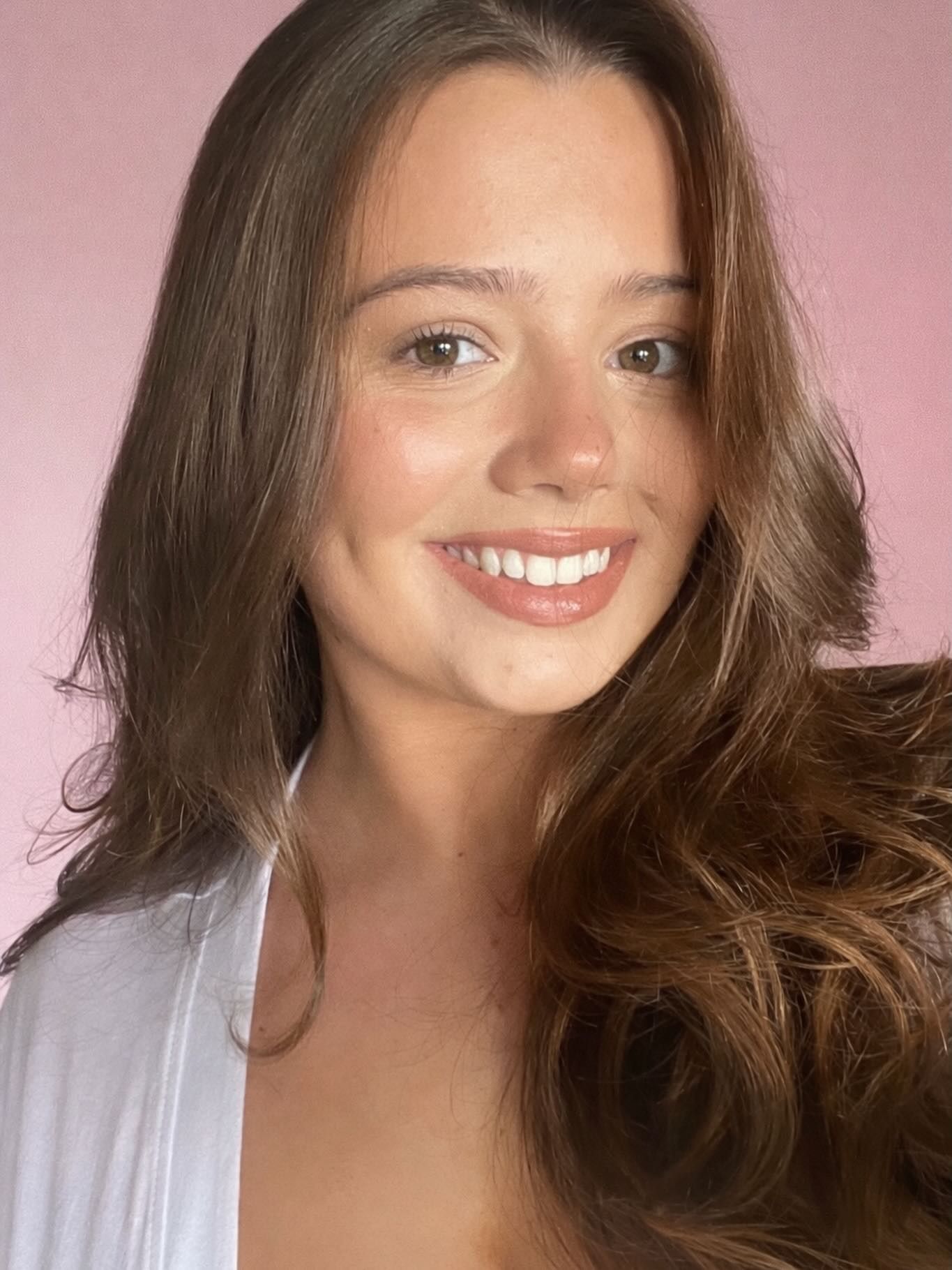 Woman with long brown hair, smiling with dimples, white top, pink background.