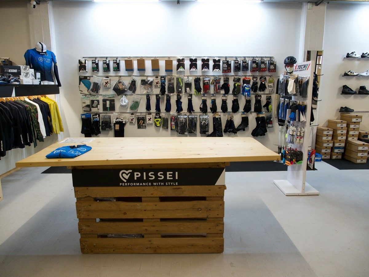 Interno di un negozio di abbigliamento ciclistico con un tavolo espositivo in legno, accessori a parete e scaffali per i vestiti.