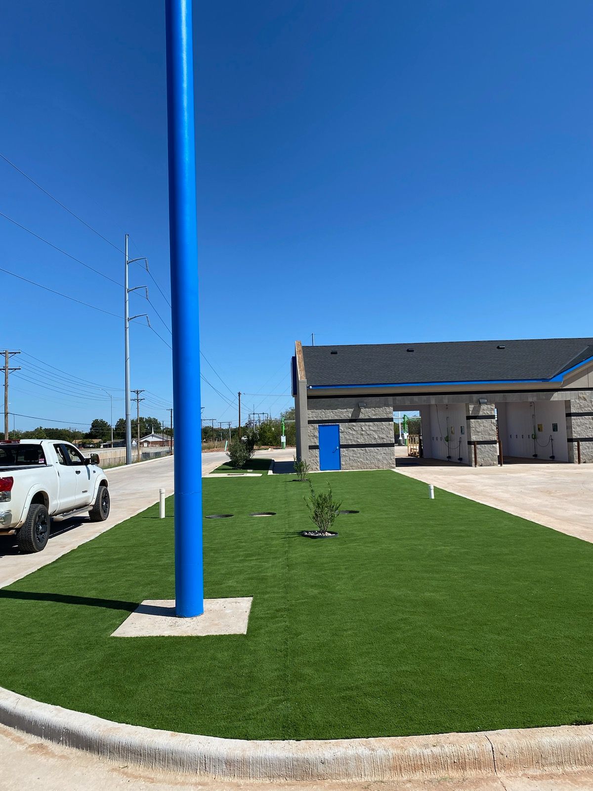 Blue flagpole on a grassy roundabout near a building and parked white truck under a clear sky