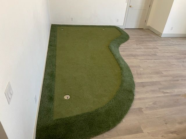 Indoor putting green in a room with wood flooring and a white wall, shaped like a golf green.