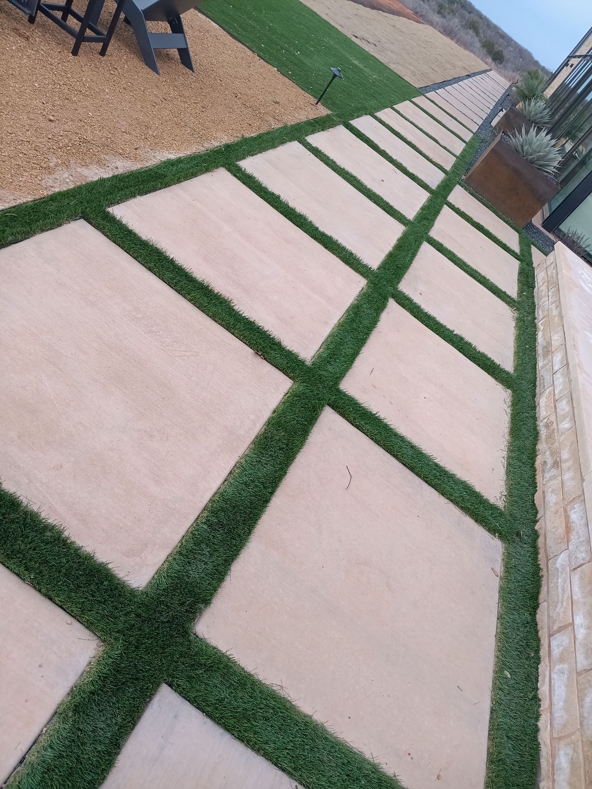 Diagonal stone walkway with green grass strips beside a gravel area and brick wall