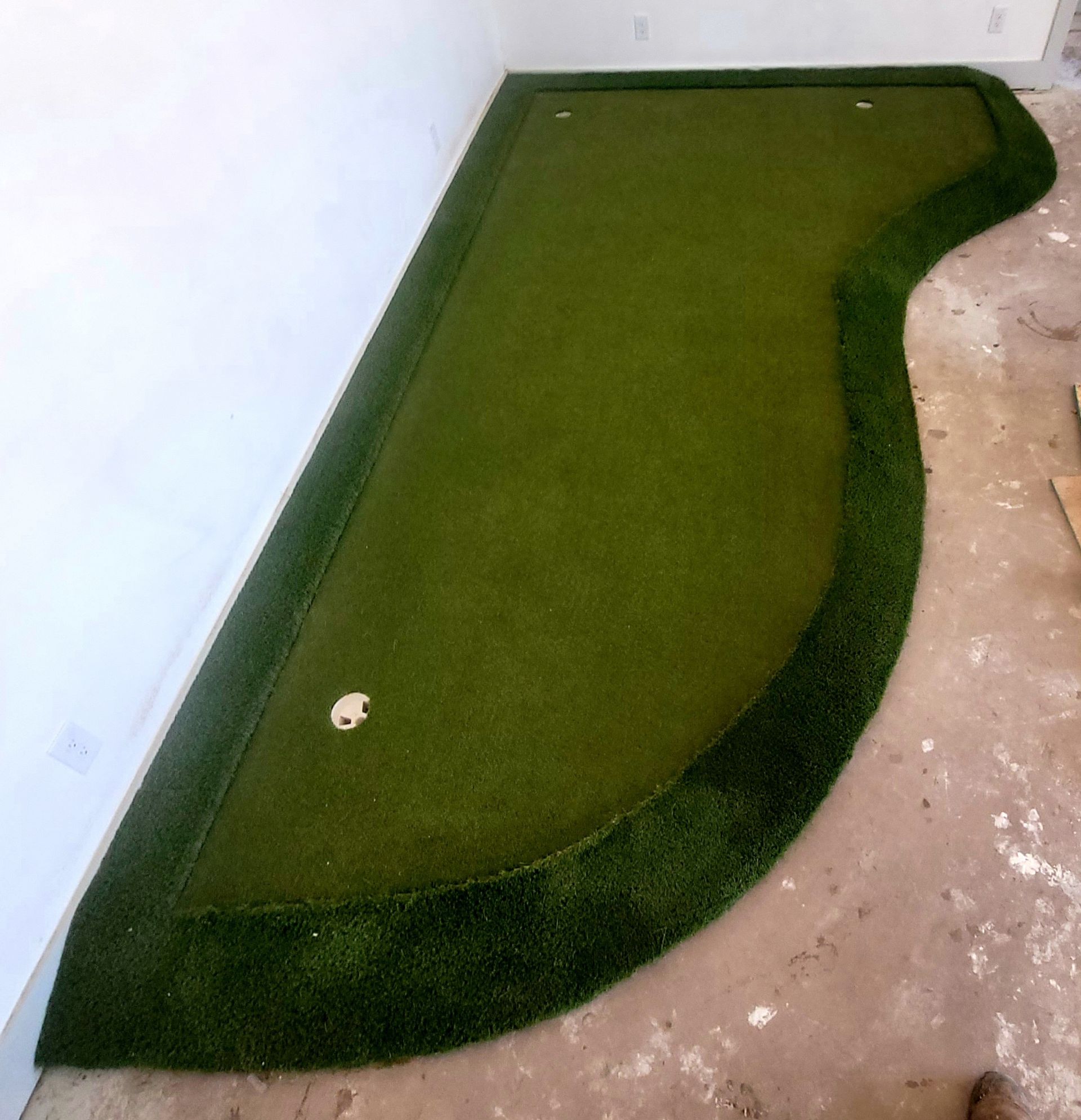 Green artificial-turf putting green in a room with a single golf hole and dark border edges