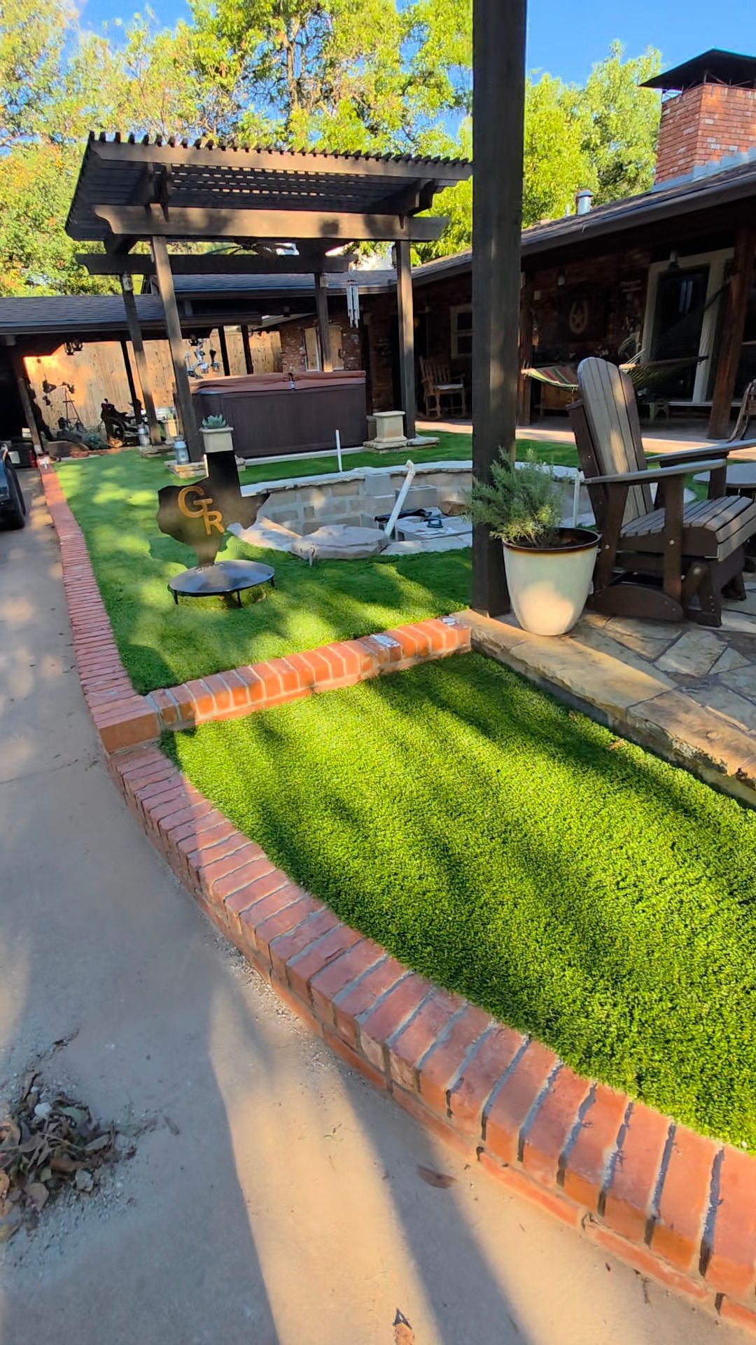 Sunny backyard patio with brick edging, green lawn, pergola, seating area, and a small pond