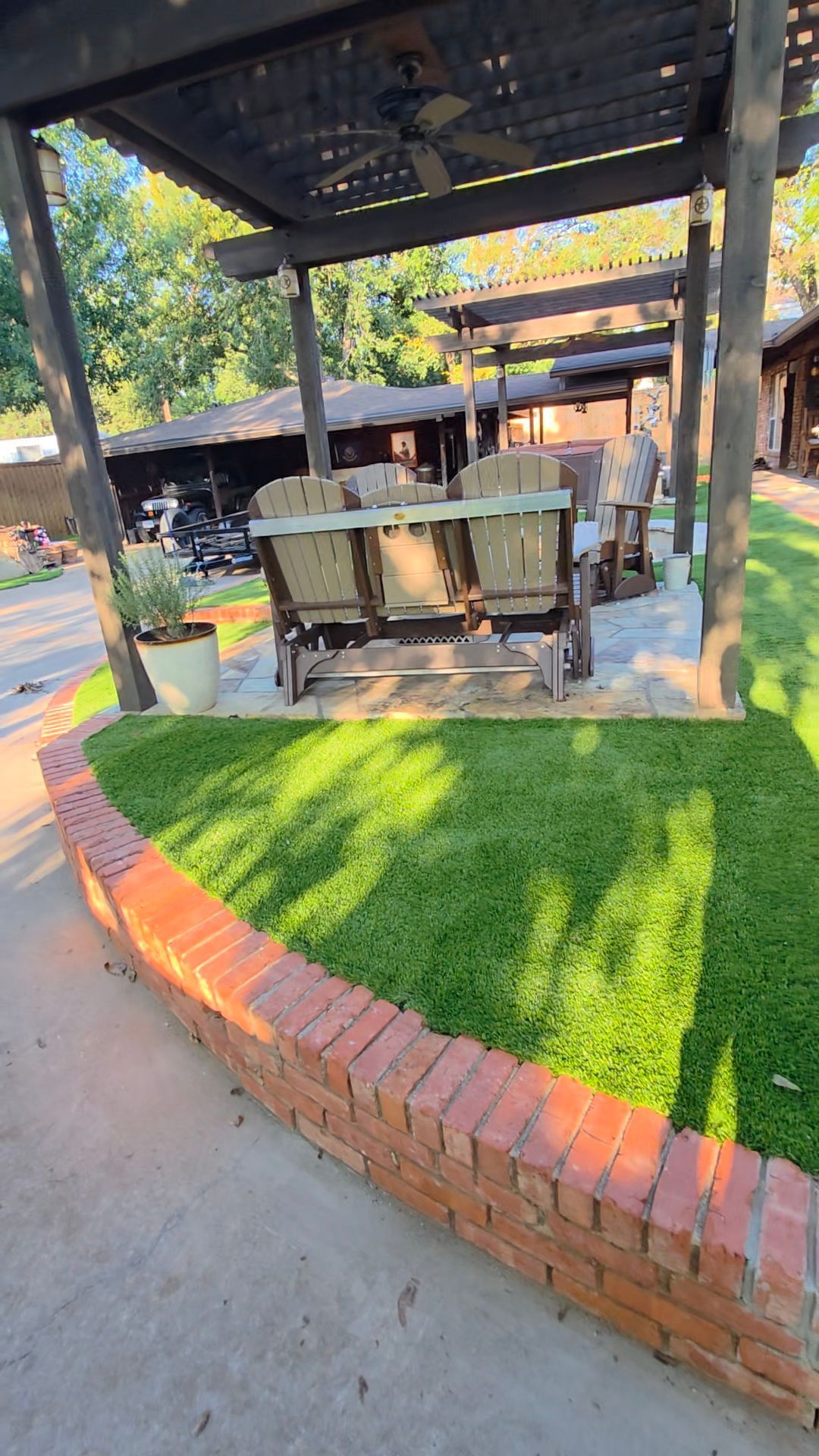 Covered outdoor seating area with tables under a metal canopy, bordered by a curved brick planter and green grass