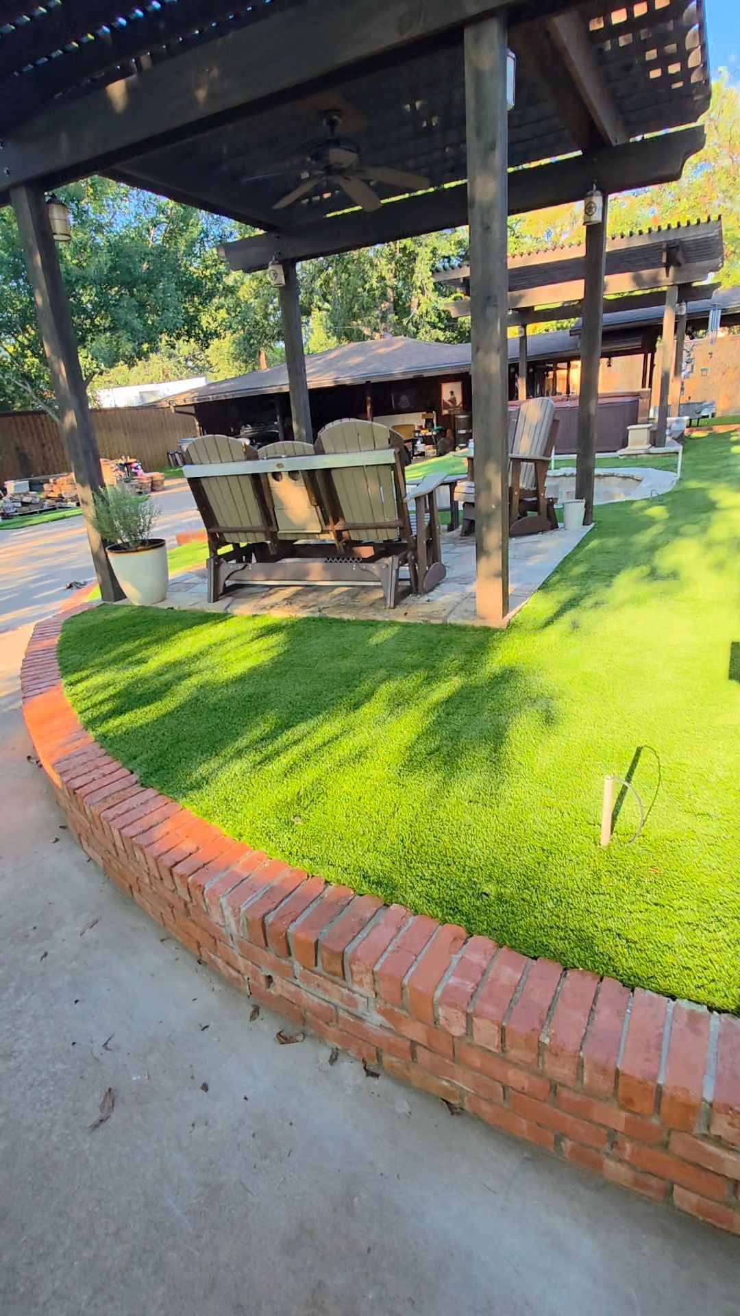 Outdoor patio with shaded seating, wooden tables, and bright green grass bordered by a brick edge.