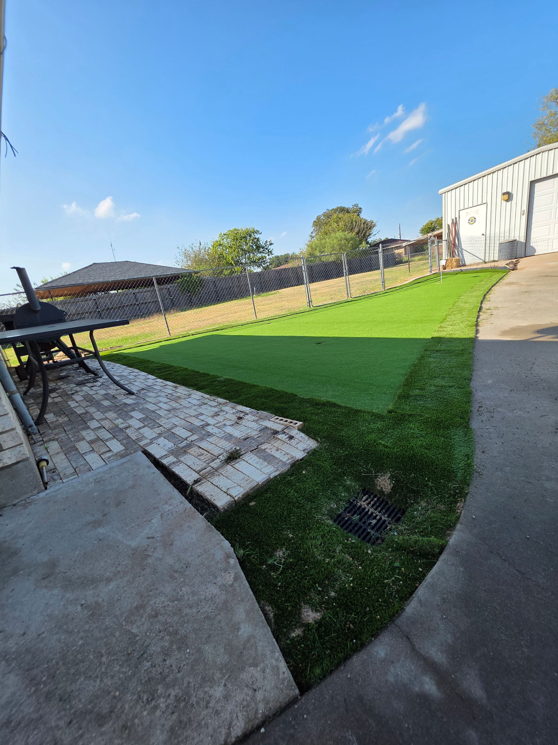Backyard patio beside bright green artificial lawn, stone path, and fence under a blue sky