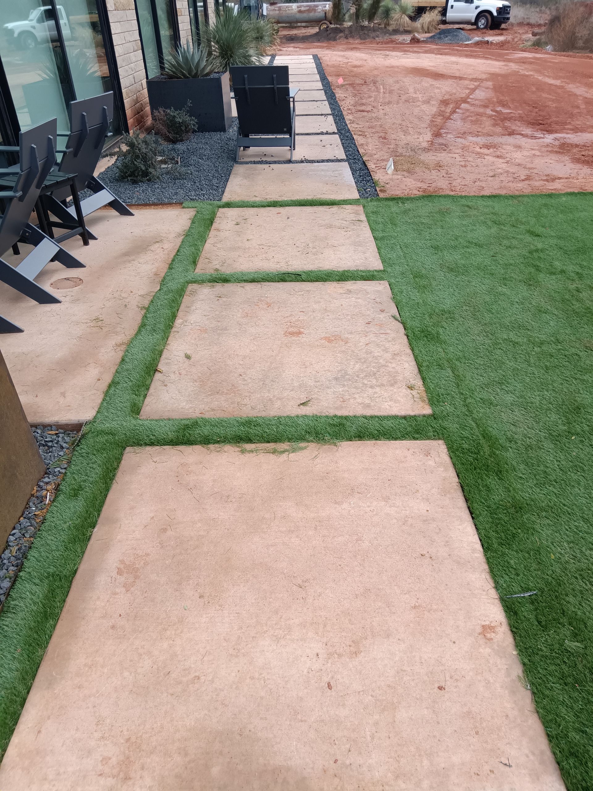 Concrete stepping-stone walkway beside a house, bordered by grass and gravel, leading toward a dirt driveway