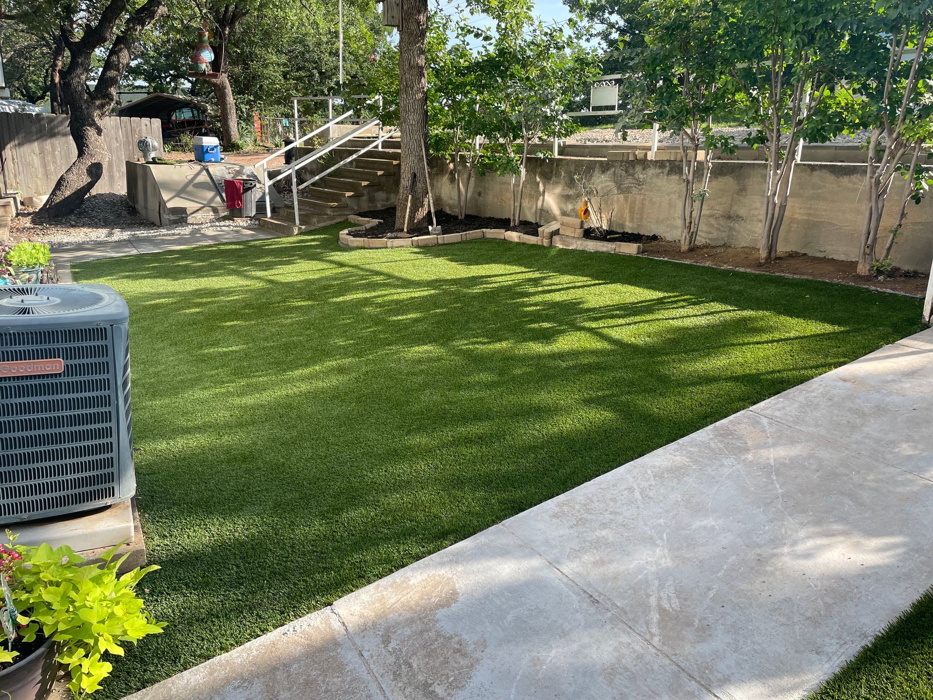 Sunny backyard with green lawn, concrete walkway, trees, and a fence along the back.