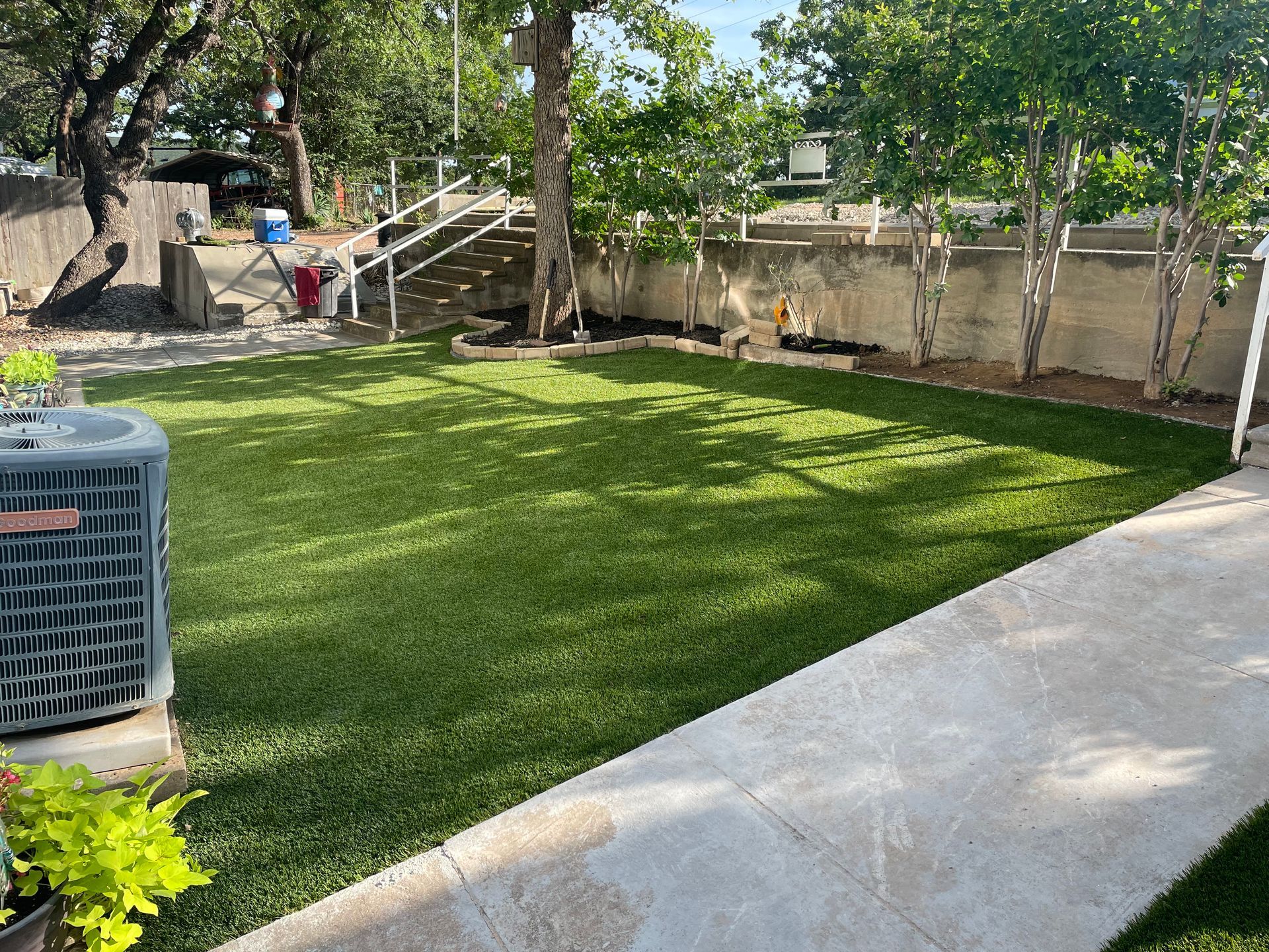 Backyard lawn with a concrete path, trees, a wall, and an air conditioner unit beside steps