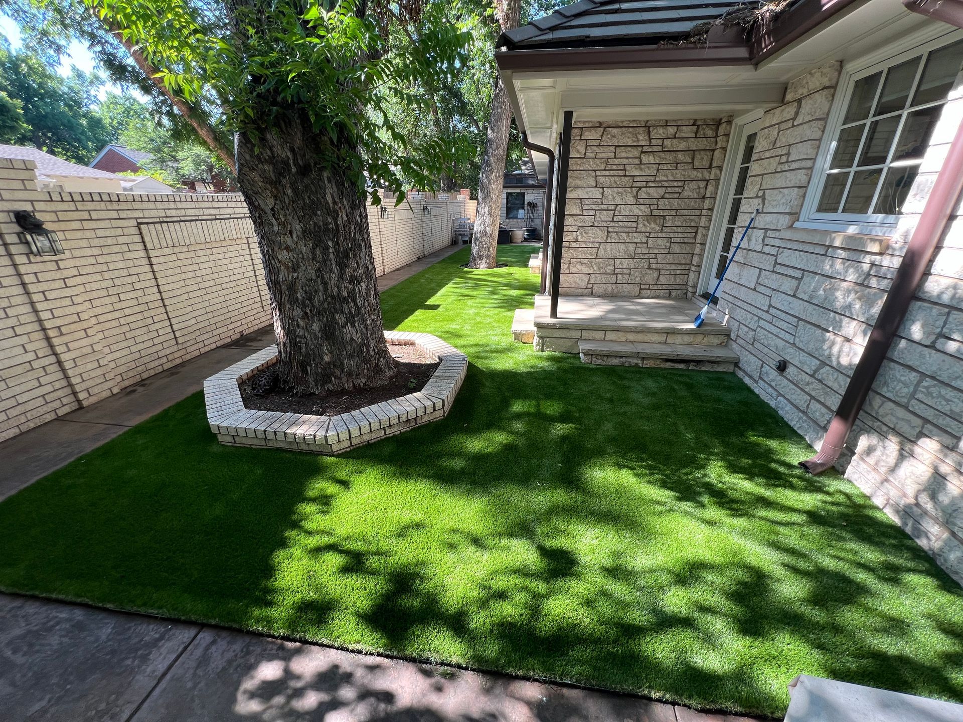 Small fenced yard with bright green artificial turf, a tree in a stone ring, and a brick house wall.
