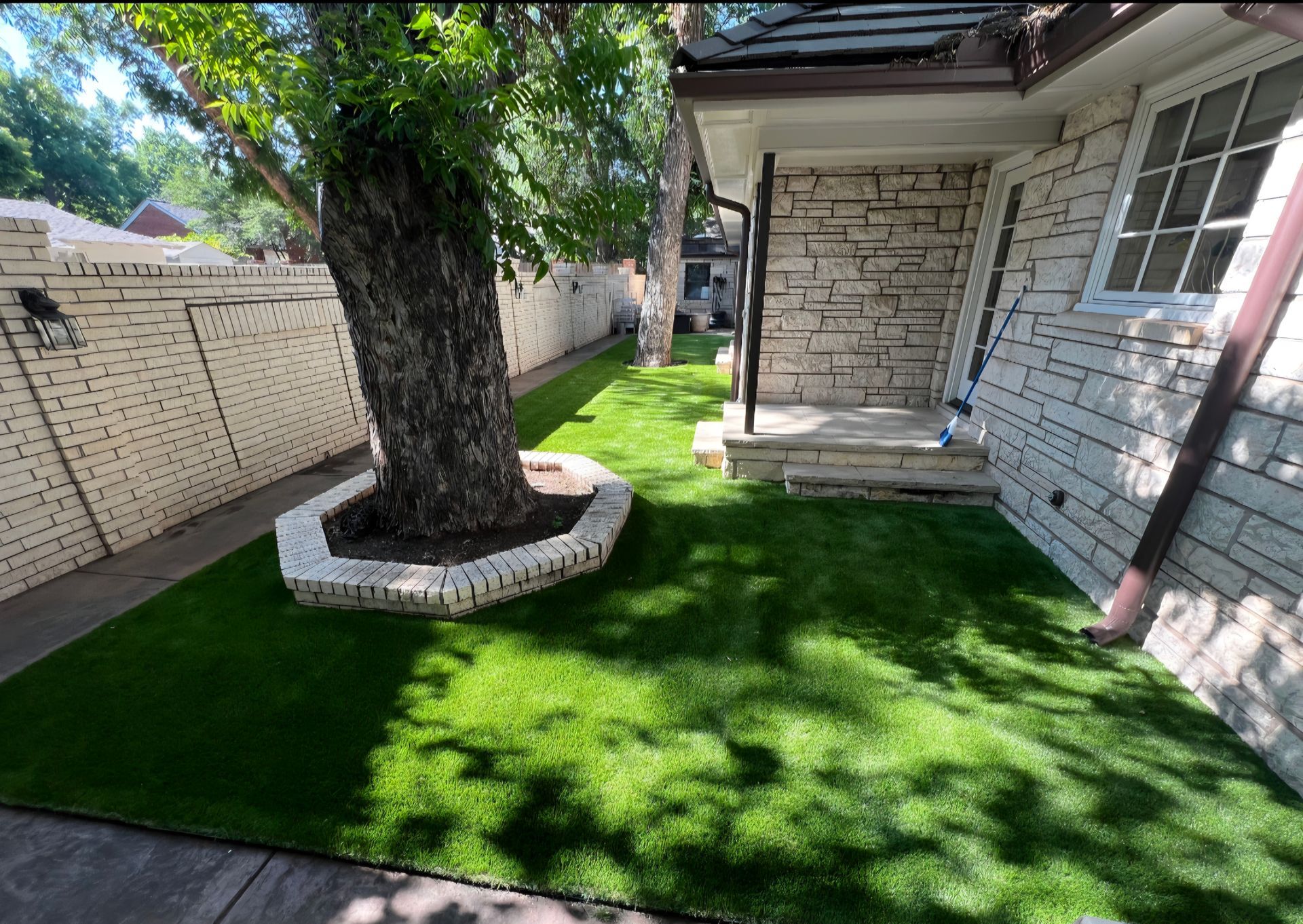 Backyard with bright green turf, a large tree, and a white house patio beside a brick wall.