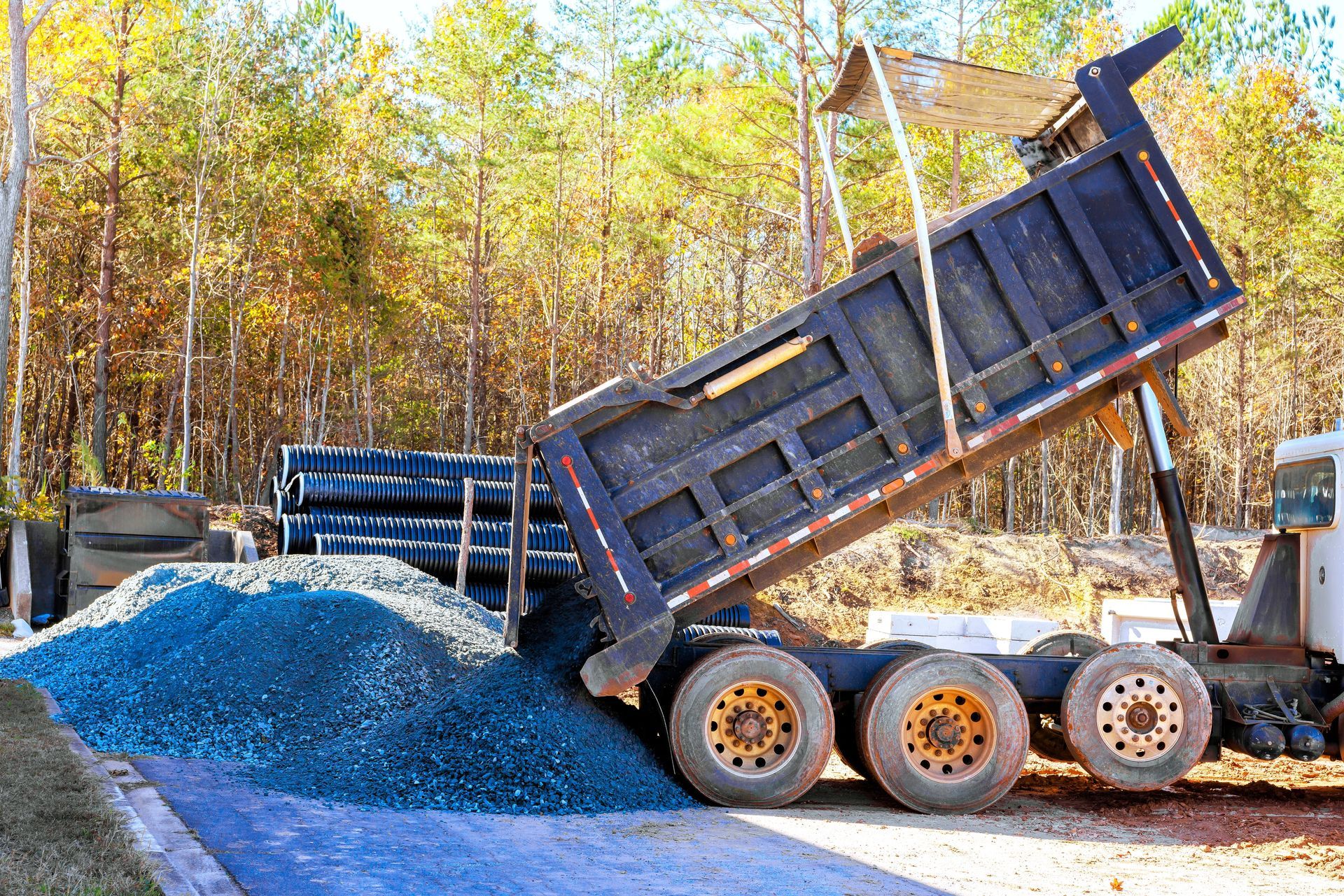 Dump truck dumping gravel onto a paved area, with pipes and trees in the background.