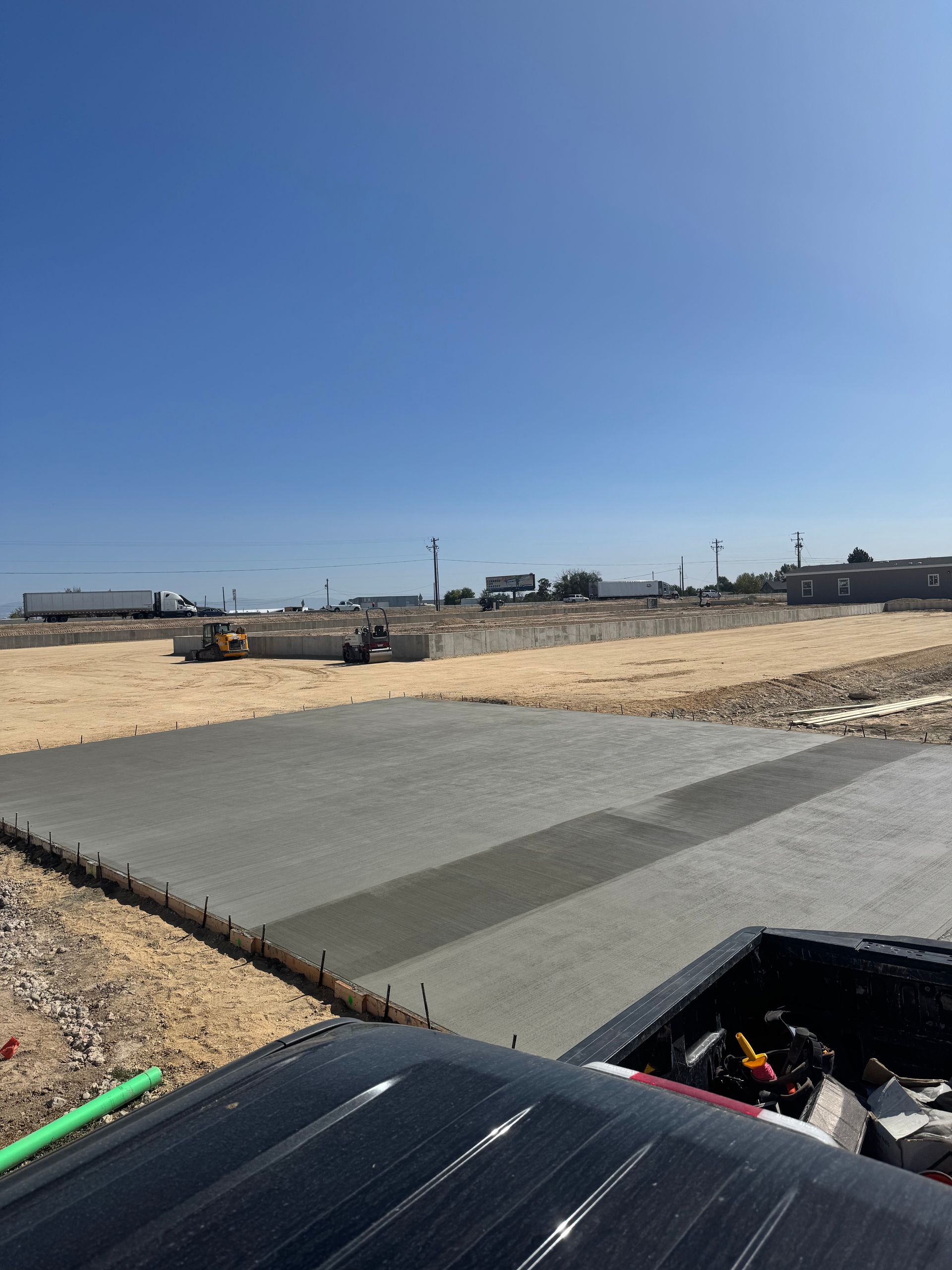 Construction site with freshly poured concrete slab; excavator in background. Clear blue sky.