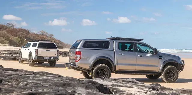 Two pickup trucks on a sandy beach. One white, one silver, both with caps. Blue sky and ocean in the background. — TJM Albury Pty Ltd in South Albury, NSW