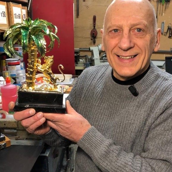 In un laboratorio, una persona sorridente con un maglione grigio tiene in mano una piccola statuetta dorata di una tigre che si arrampica su una palma.