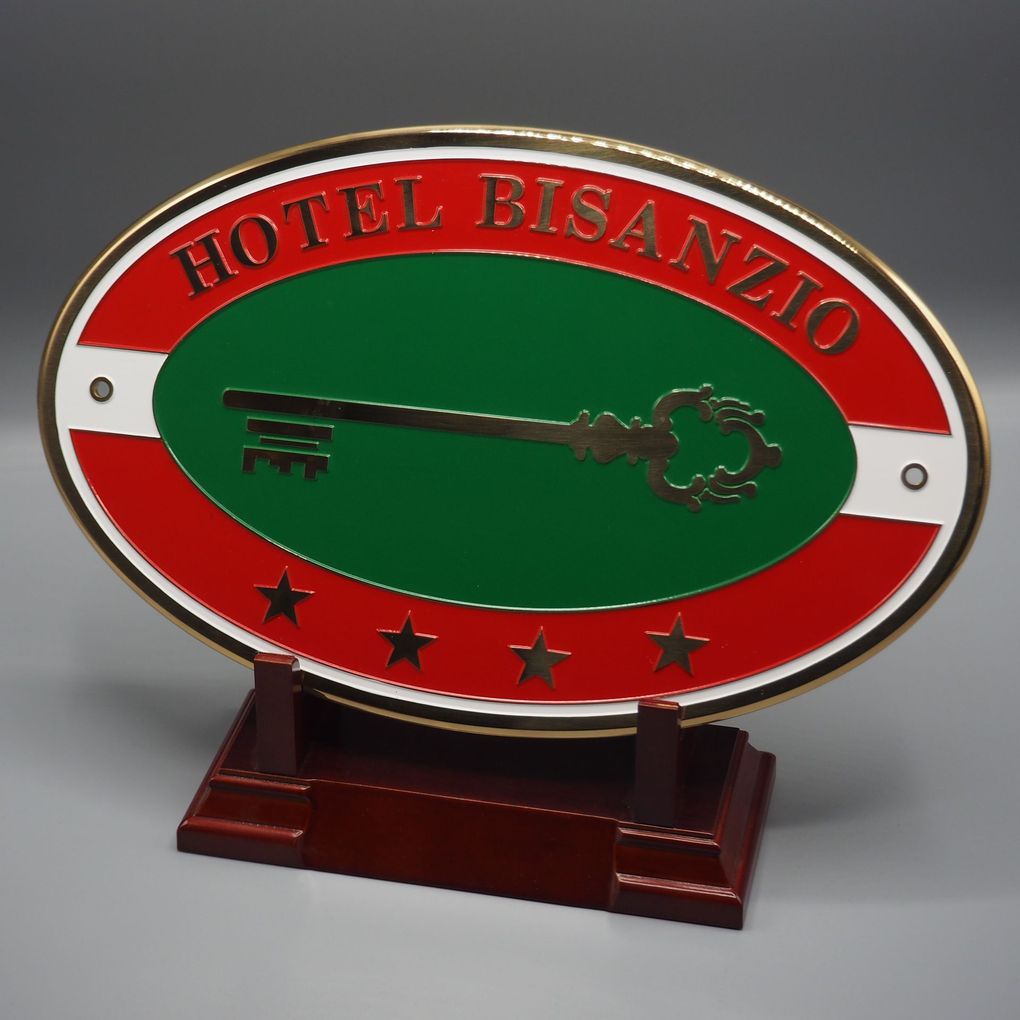 Insegna ovale dell'Hotel Bisanzio con l'icona di una chiave, quattro stelle dorate e colori rosso, verde e bianco su un supporto in legno.