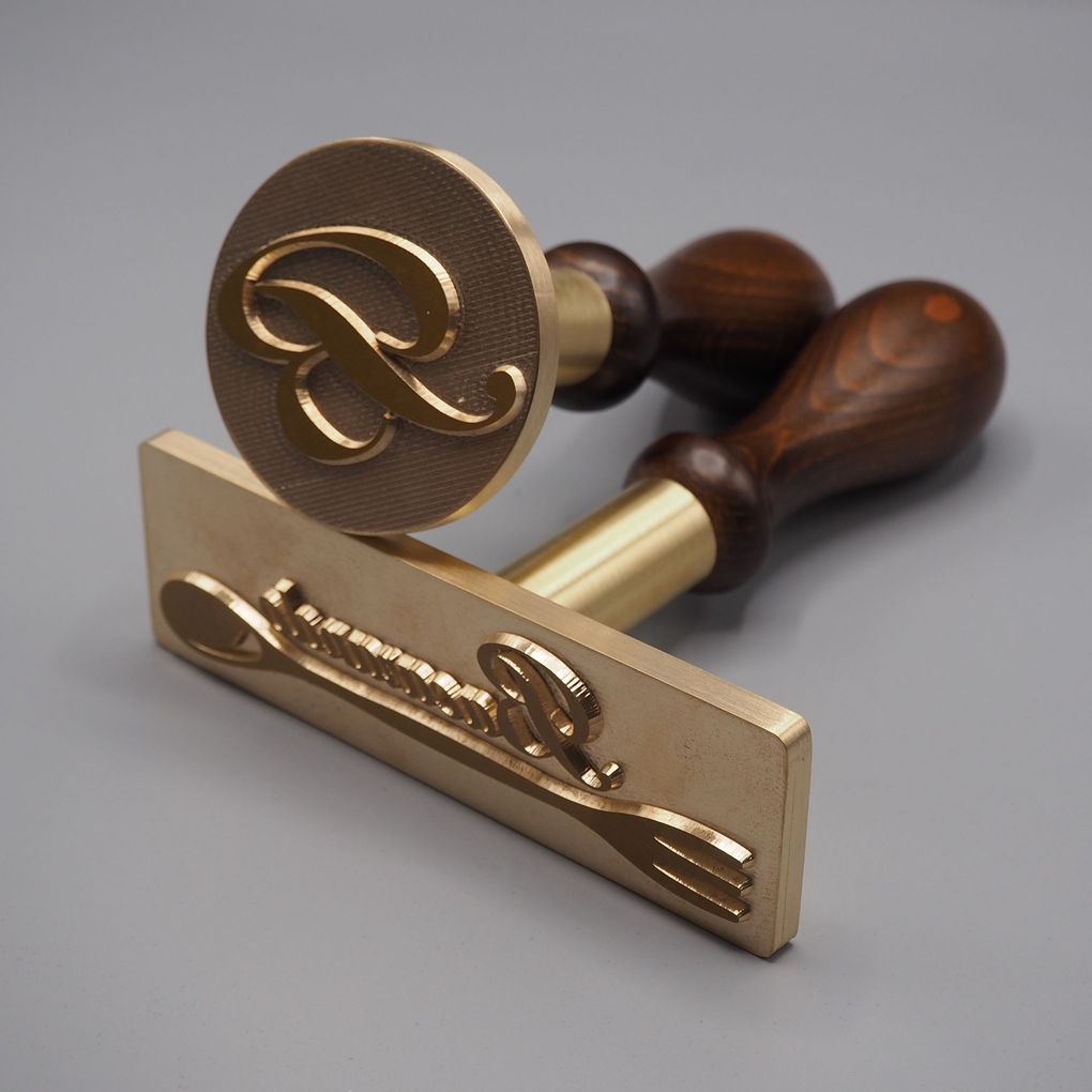 Due marchi a fuoco in ottone con manici in legno, uno circolare con una "R" e uno rettangolare con una forchetta e il logo "R".