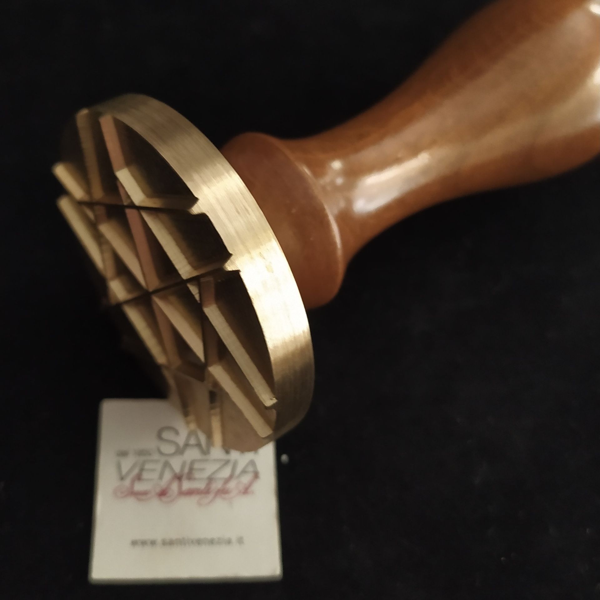 Un timbro in ottone con motivo a griglia, fissato a un manico di legno, sospeso sopra un cartoncino con il logo del marchio su una superficie nera.