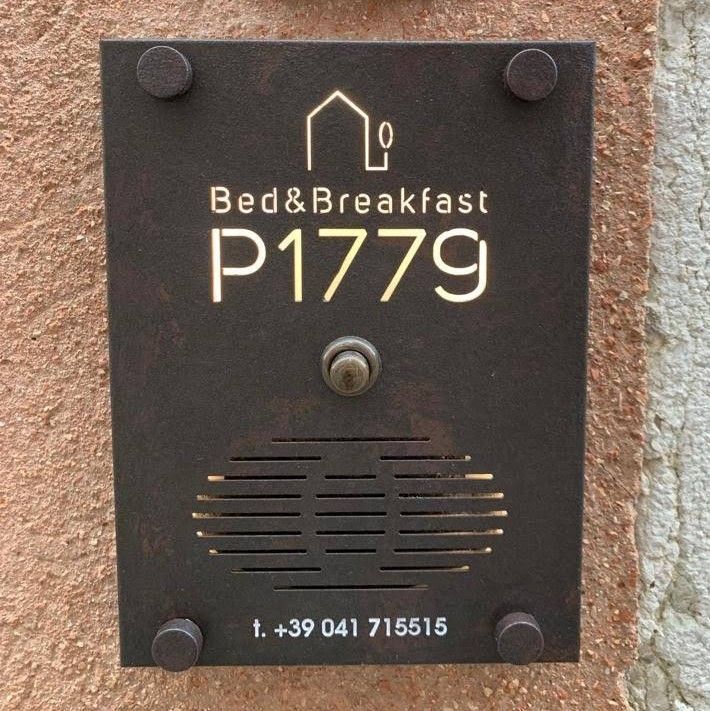 Pannello citofonico in metallo scuro per Bed & Breakfast P1779 con numero di telefono e icona di casa, montato su una parete con finitura materica.