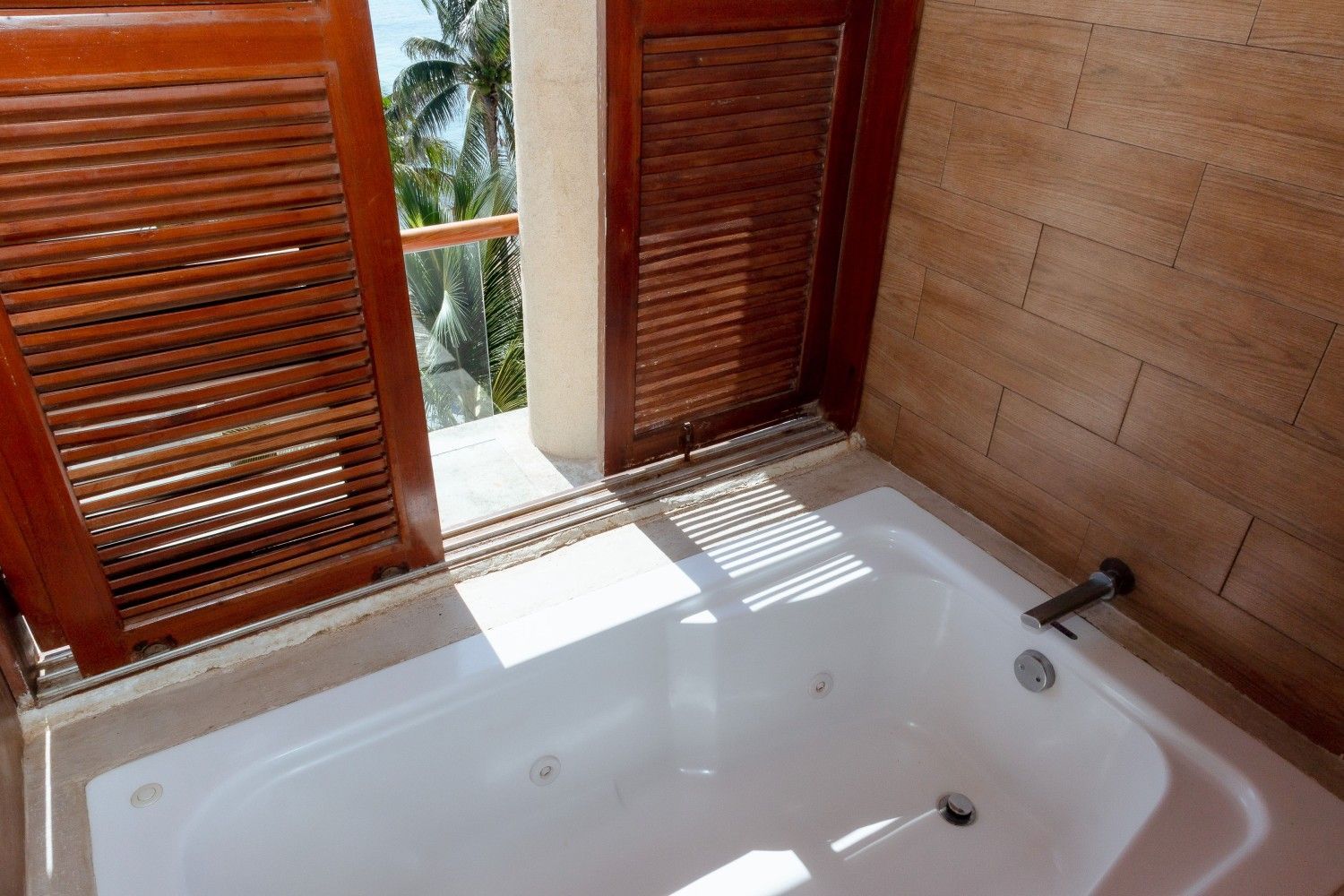 Bañera blanca junto a ventanas con celosías de madera, con la luz del sol proyectando sombras en el interior.