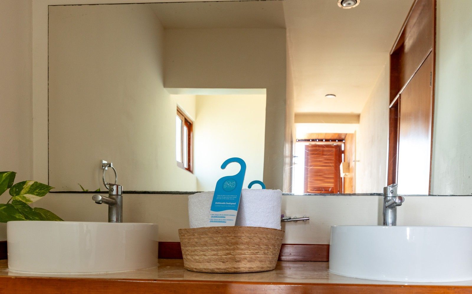 Mueble de baño con dos lavabos blancos, una cesta de mimbre, espejo y toallas en una habitación luminosa y moderna.
