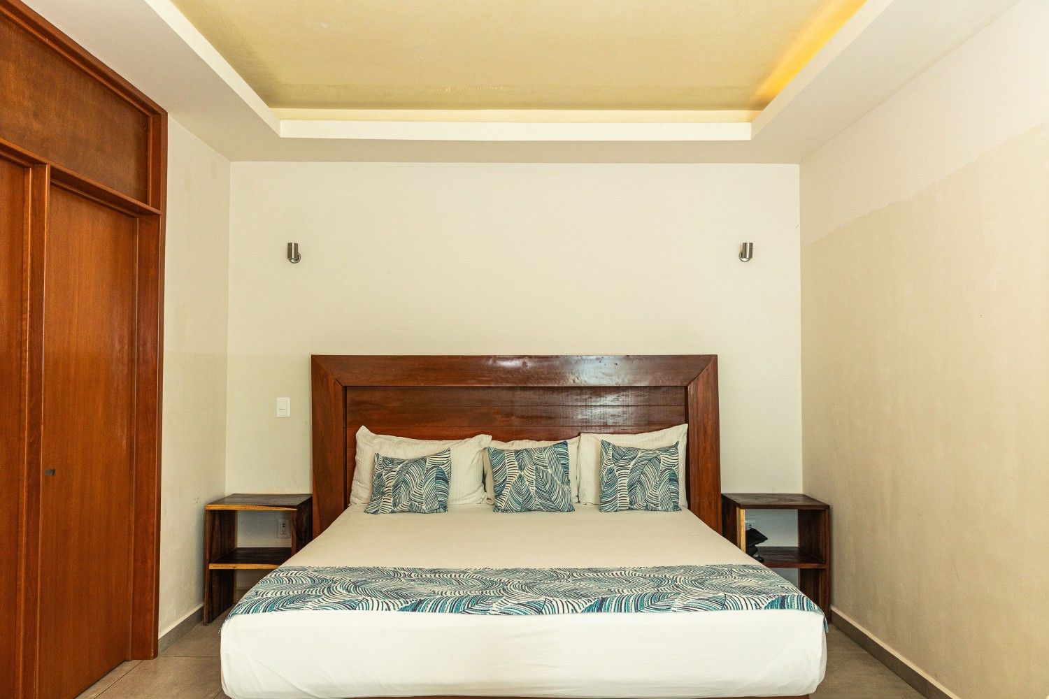 Dormitorio minimalista con cama de madera, almohadas estampadas y mesitas de noche en una habitación de color crema.