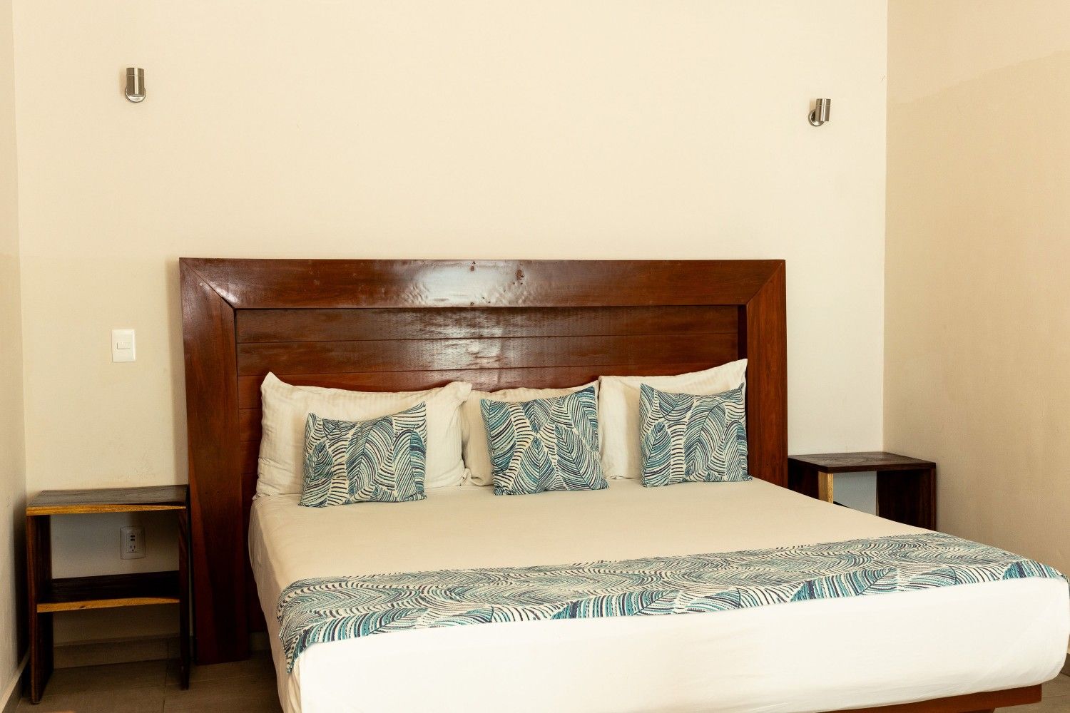 Dormitorio con cama de madera, ropa de cama blanca y dos mesitas de noche oscuras sobre paredes beige.