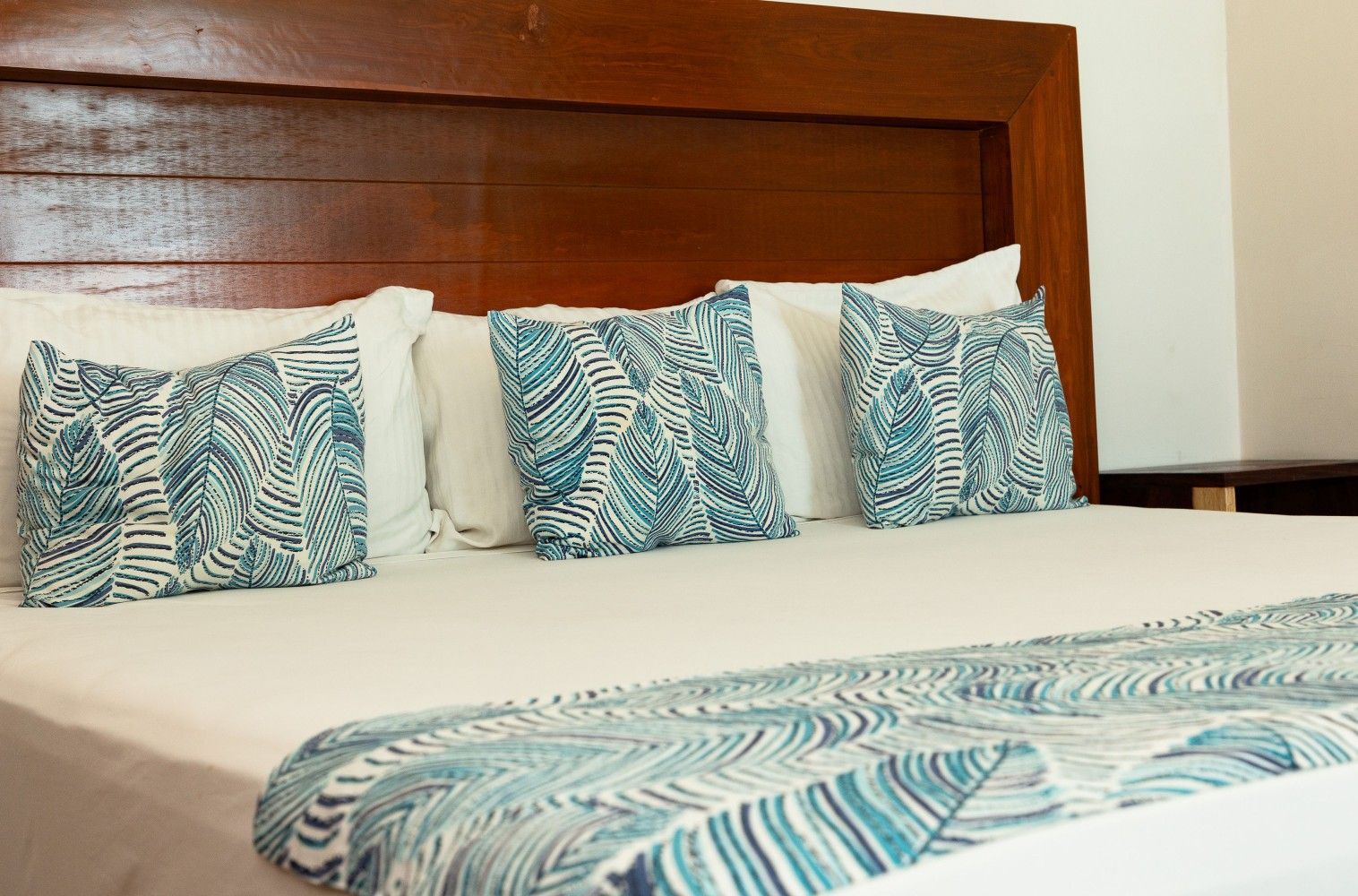 Cama con sábanas blancas, cojines de hojas azules y camino a juego, con cabecero de madera.