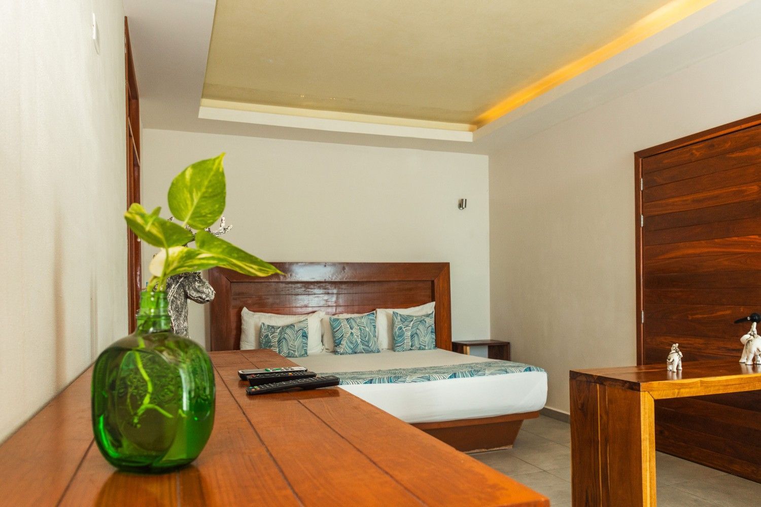 Dormitorio moderno con cama de madera, ropa de cama blanca, jarrón verde sobre la mesa e iluminación cálida en el techo.