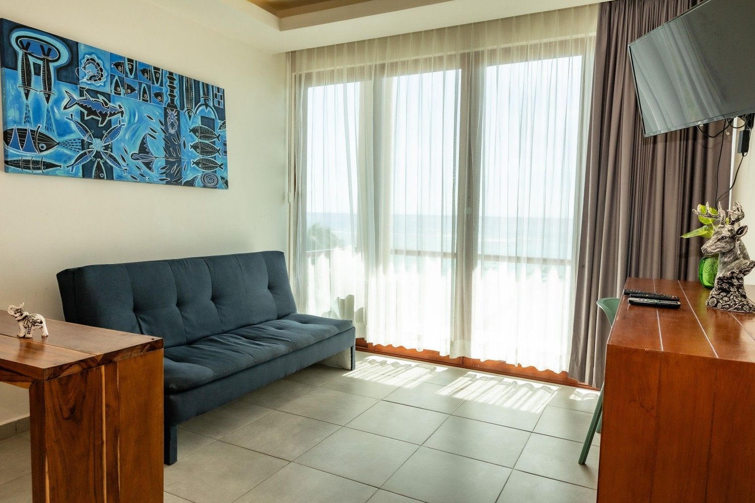 Luminoso salón con sofá azul, armarios de madera, cuadros en las paredes y grandes ventanales con cortinas transparentes.