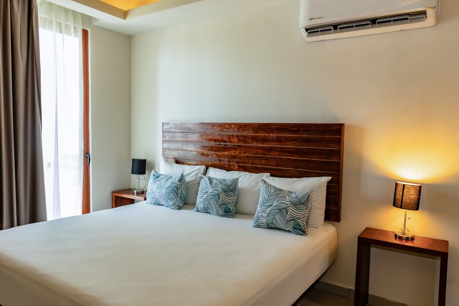 Habitación luminosa con una cama grande, cabecero de madera, lámparas de noche y aire acondicionado cerca de una ventana.