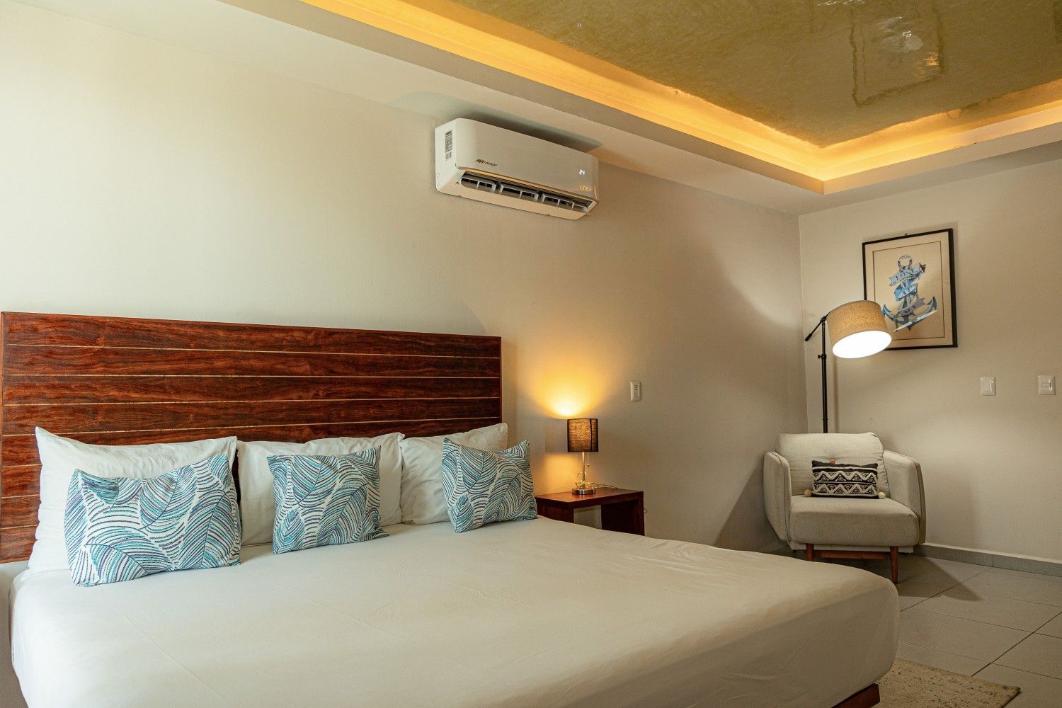 Habitación de hotel moderna con cama king size, dos lámparas, sillón, cuadro en la pared e iluminación cálida en el techo.