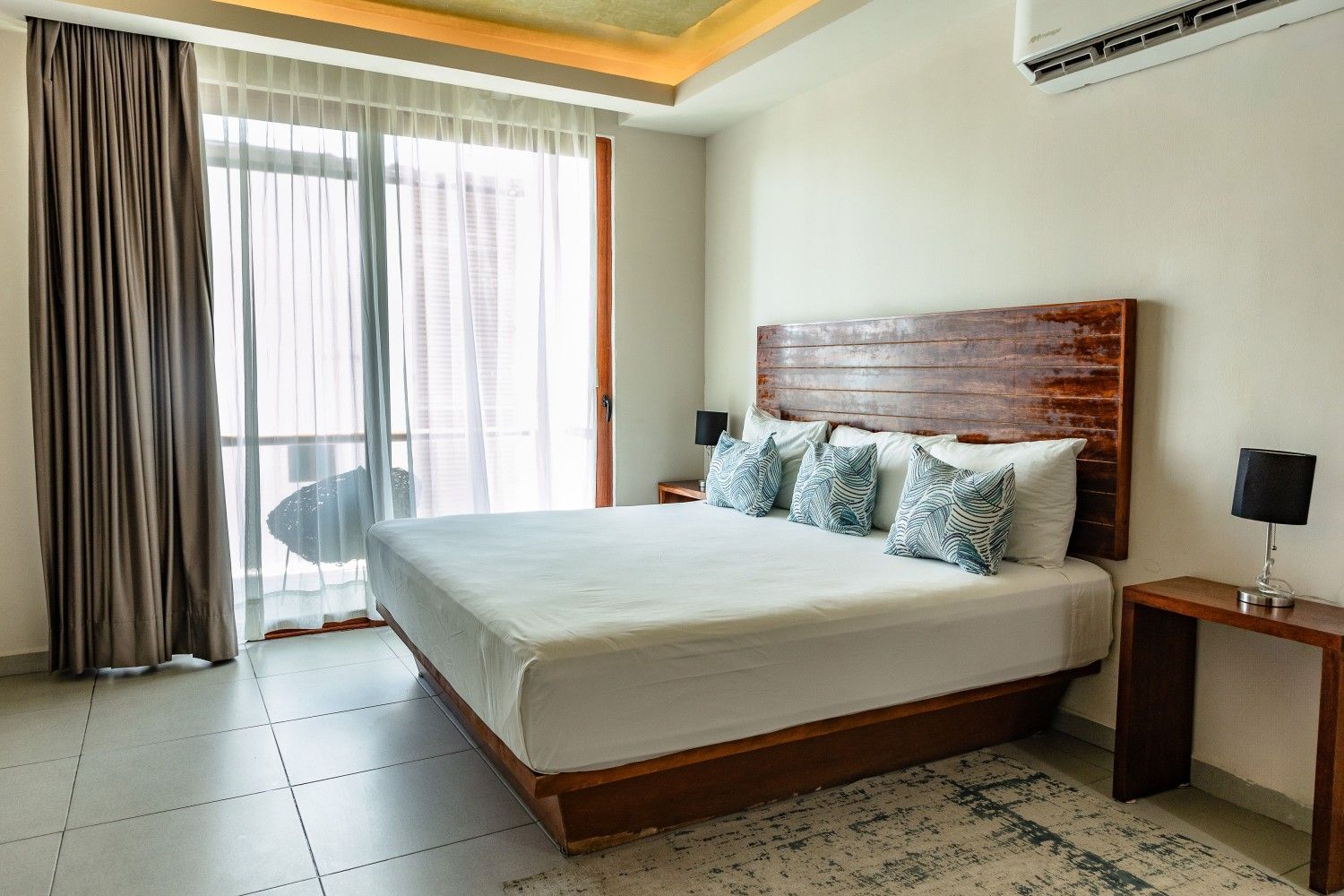 Dormitorio moderno con cama king size, cabecero de madera, mesita de noche, cortinas y puerta al balcón.