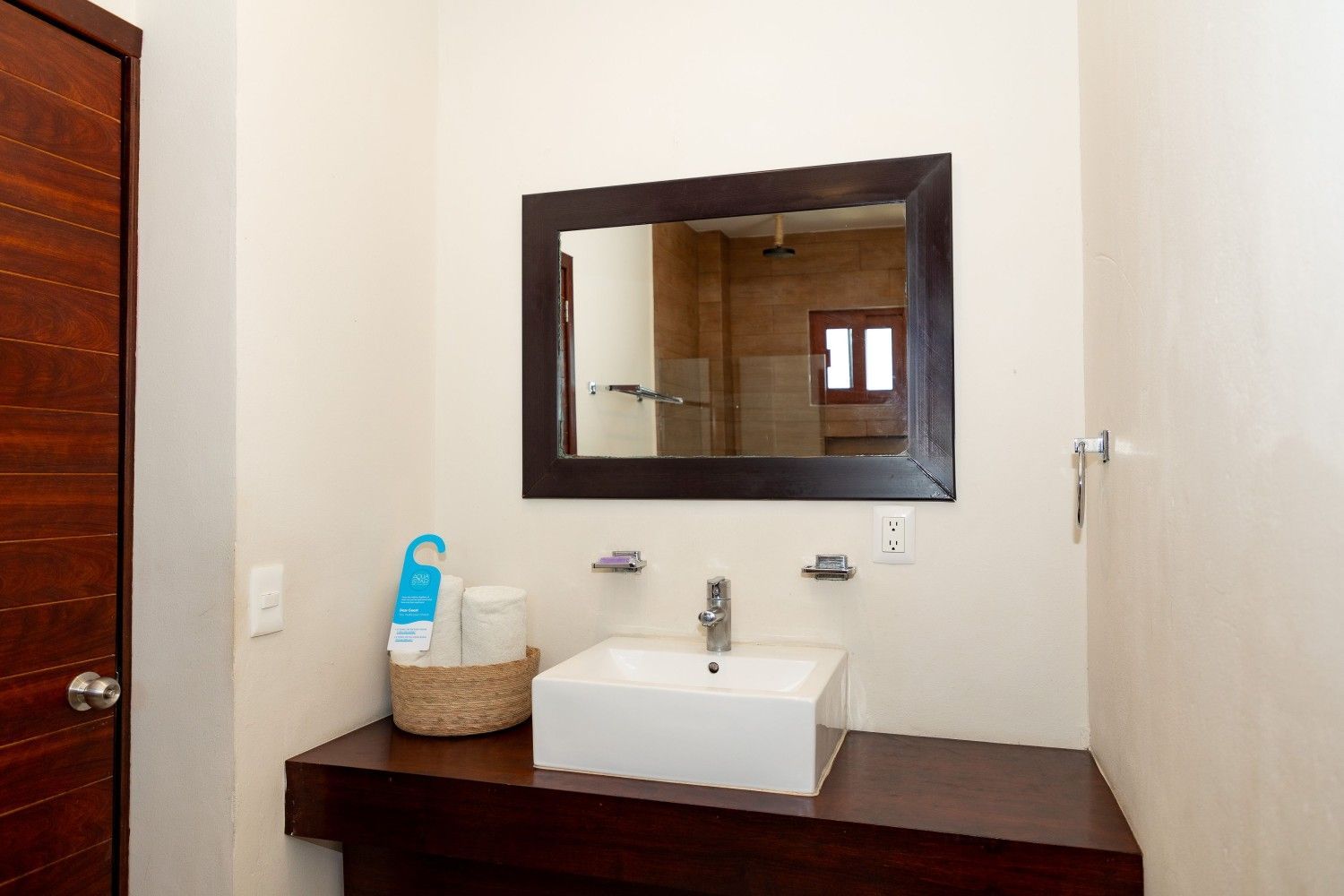 Lavabo de baño con encimera oscura y espejo enmarcado en una pared clara, con jabón y una cesta al lado.