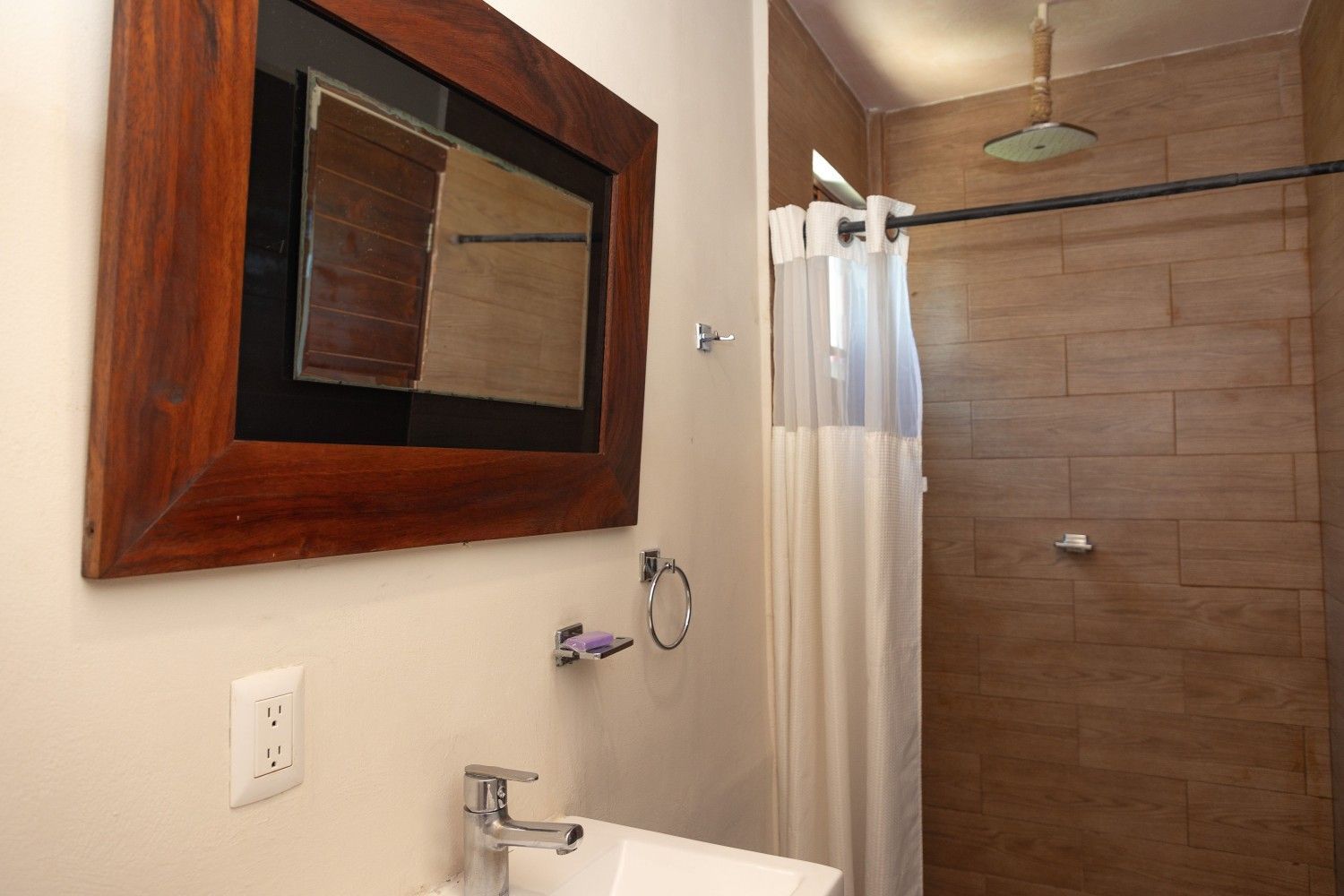 Baño con ducha de azulejos color beige, cortina blanca, lavabo y espejo grande con marco de madera.