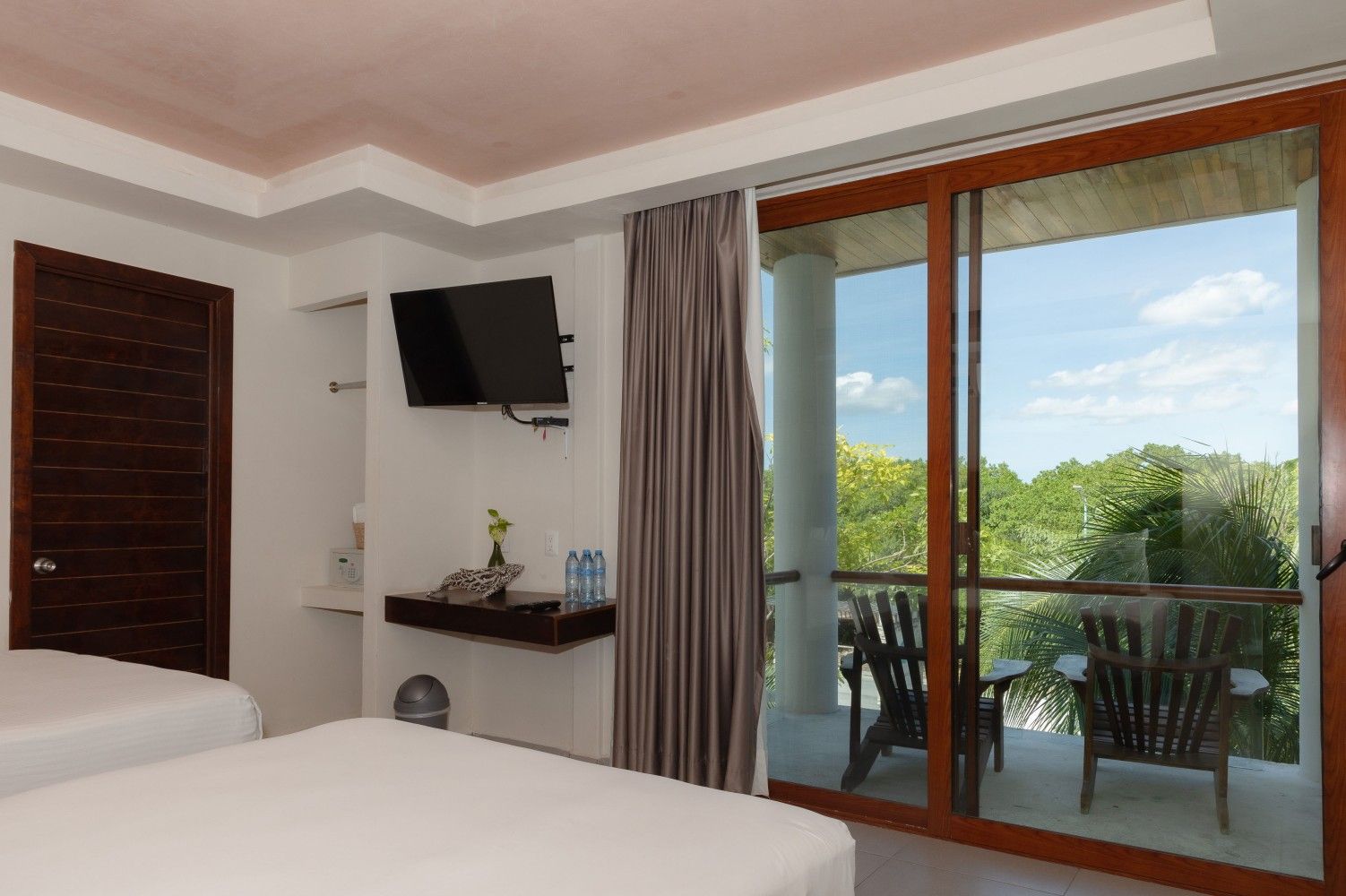 Habitación de hotel con cama, televisor de pared, escritorio y balcón con vistas a la vegetación.