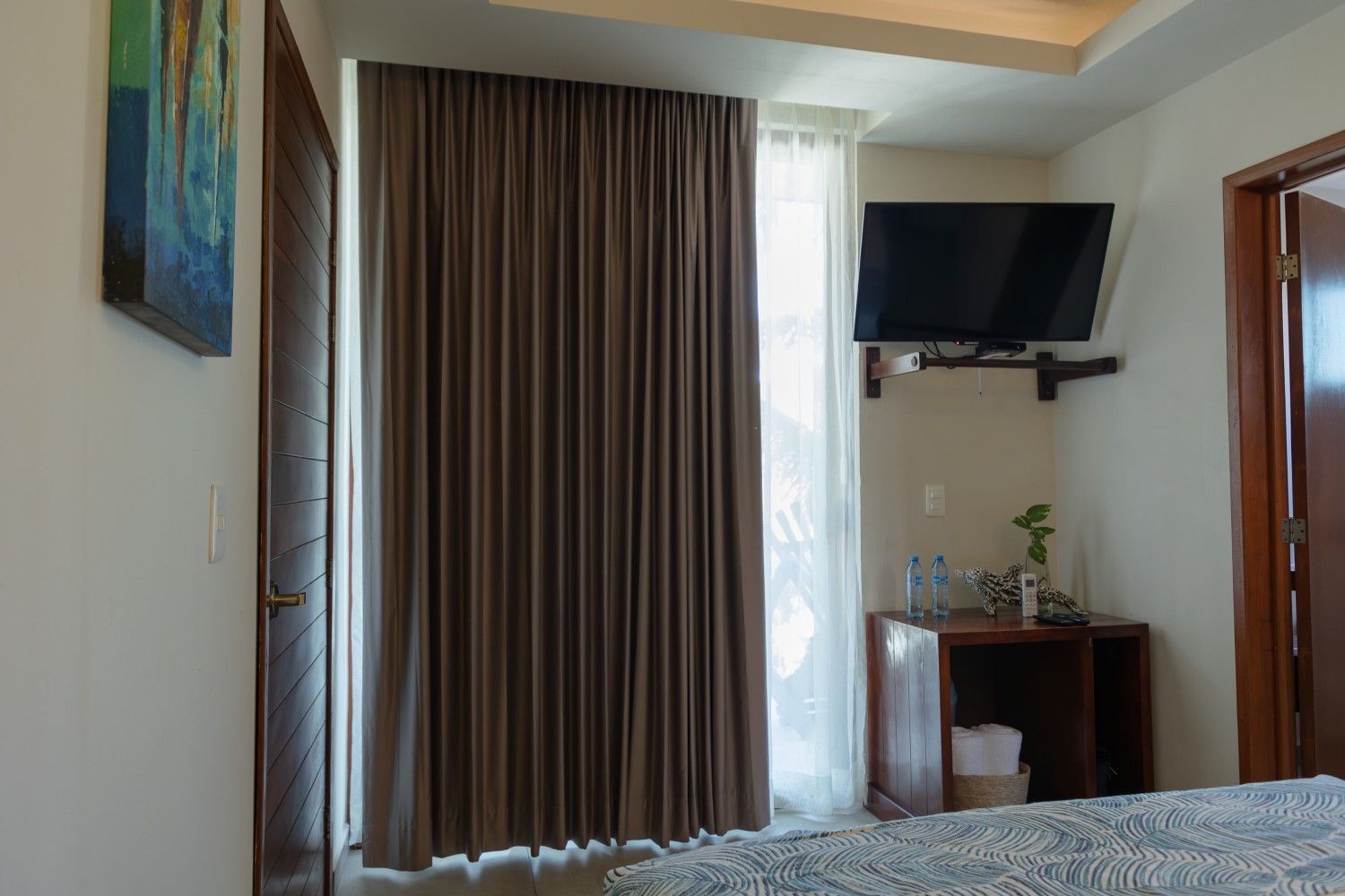 Habitación de hotel con cama, televisor de pared, escritorio, cortinas y ventana con luz natural.