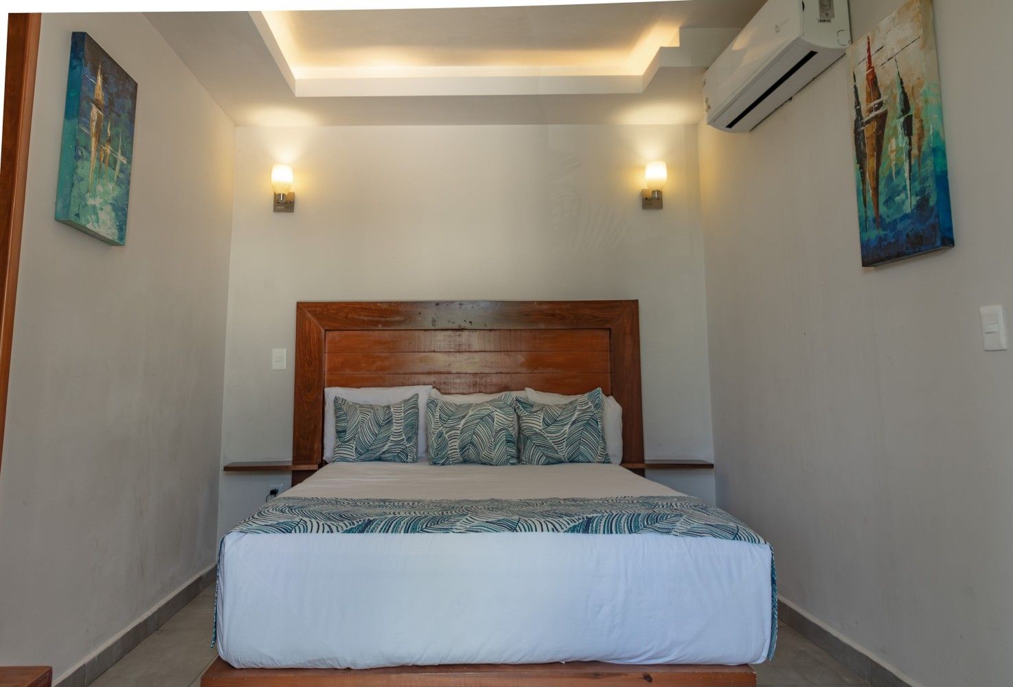 Dormitorio minimalista con cama de madera, sábanas blancas, cuadros en la pared e iluminación tenue en el techo.